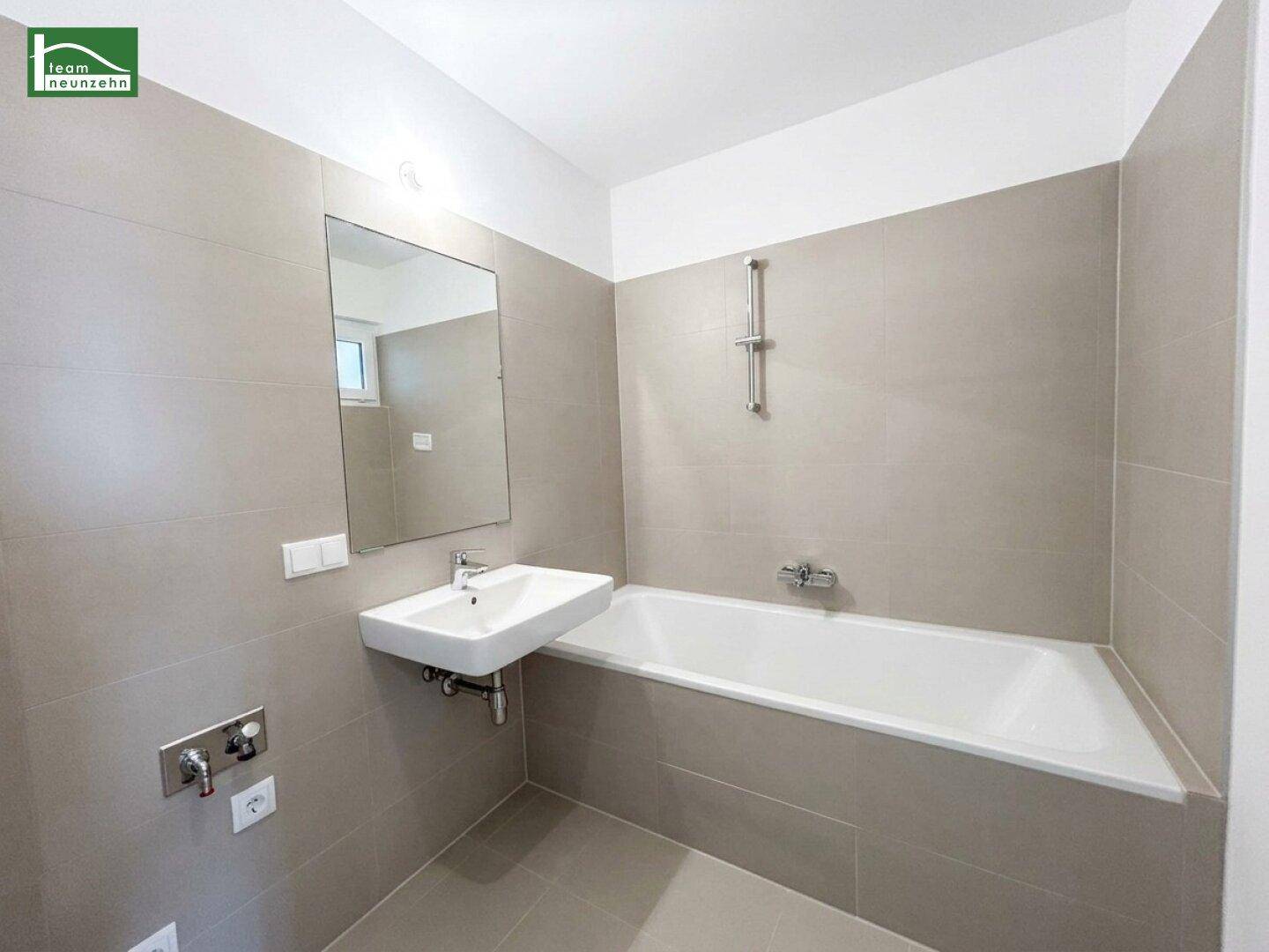 Modernes Badezimmer mit grauen Fliesen, Badewanne, Waschbecken und einem Fenster für Tageslicht.