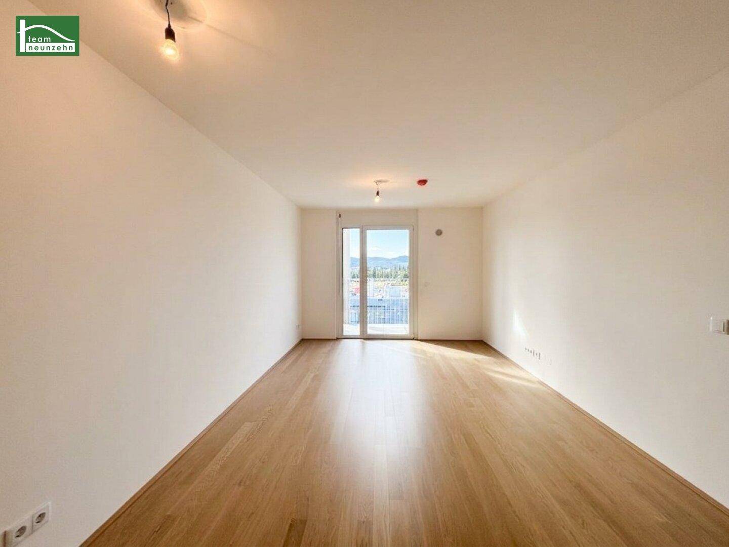 Helles, leeres Wohnzimmer mit hellem Holzfußboden und Zugang zu einem Balkon.