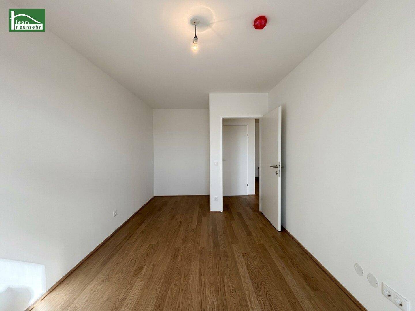 Geräumiger, heller Raum mit dunklem Holzfußboden und offener Tür zu einem weiteren Zimmer.