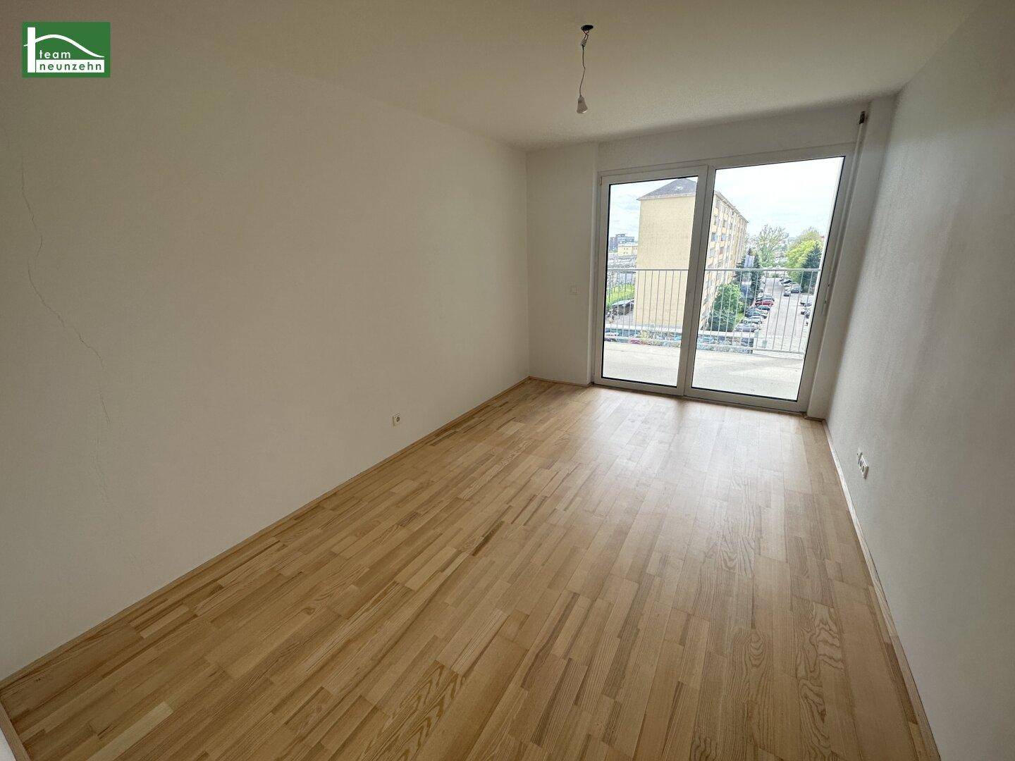 Leeres Zimmer mit Parkettboden und großen Fenstern, die viel Tageslicht hereinlassen.