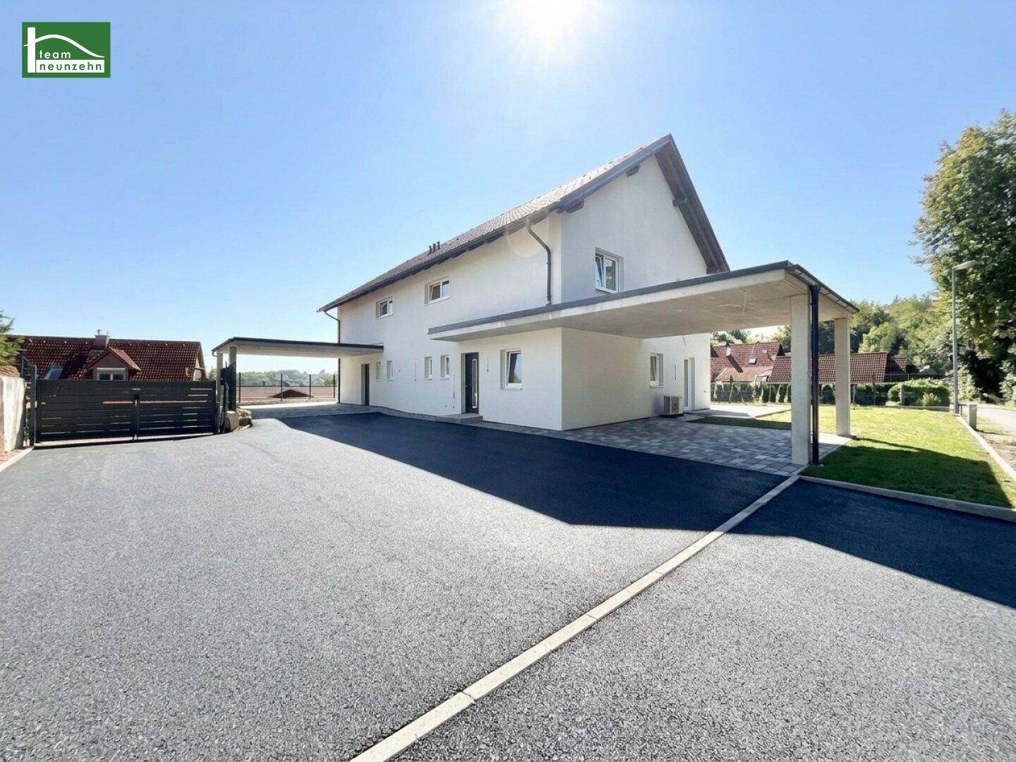 Modernes Einfamilienhaus mit heller Fassade, großem Vorplatz und zwei Carports unter strahlendem Sonnenschein.