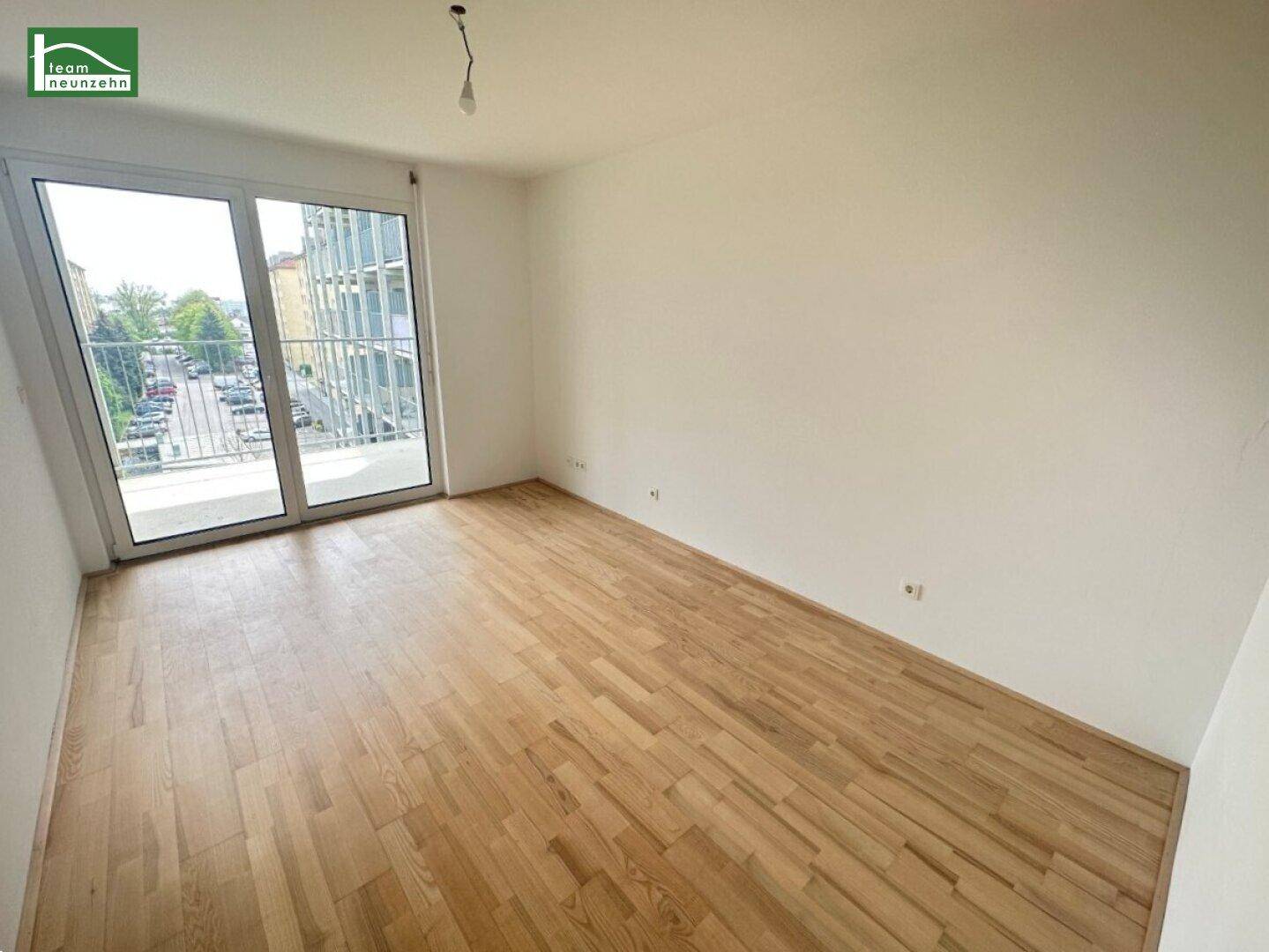 Geräumiges, helles Zimmer mit Holzboden und großen Fenstern, die viel Tageslicht hereinlassen.