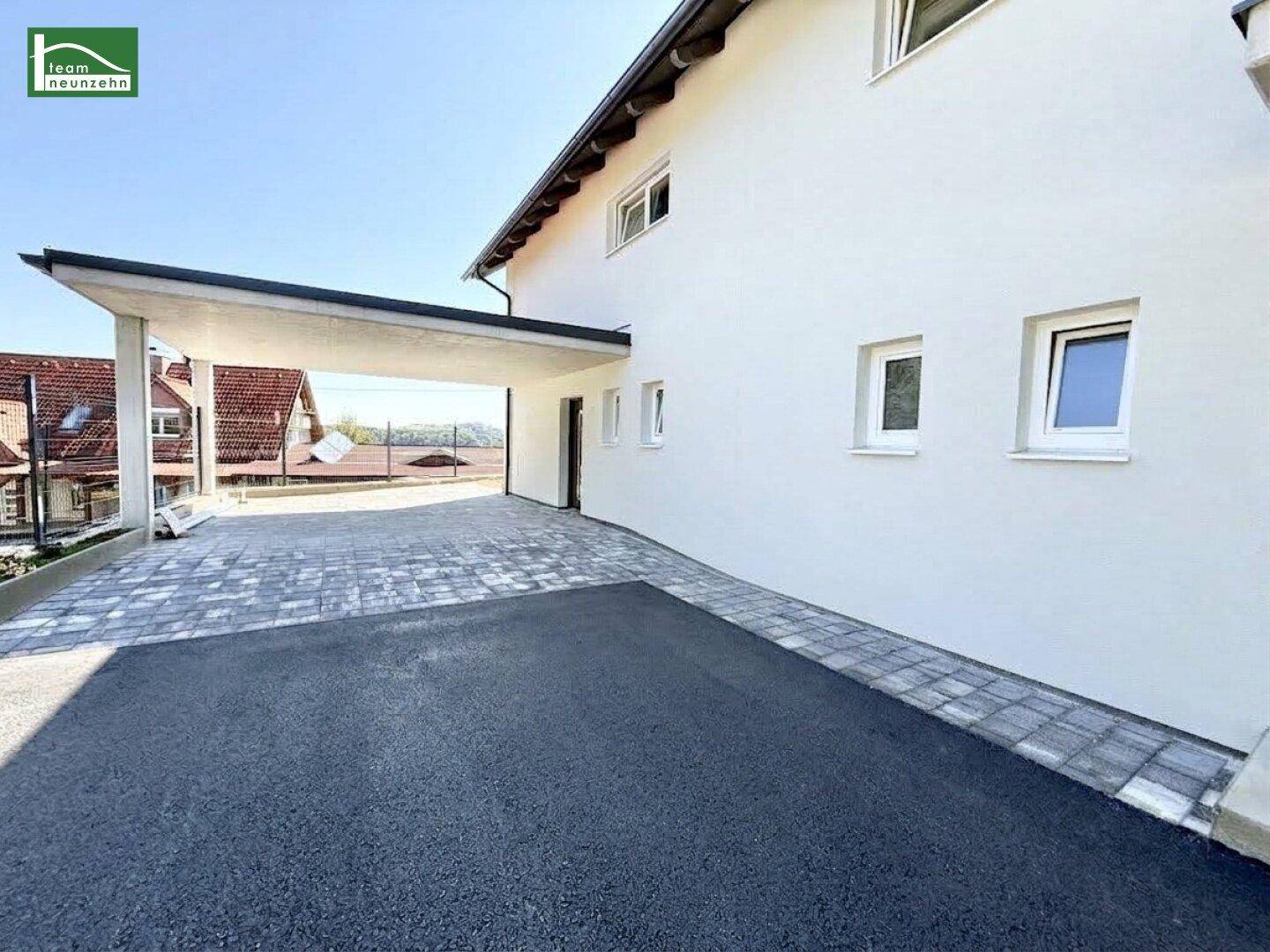 Seitenansicht des Neubaus mit Carport und gepflastertem Bereich unter klarem Himmel.