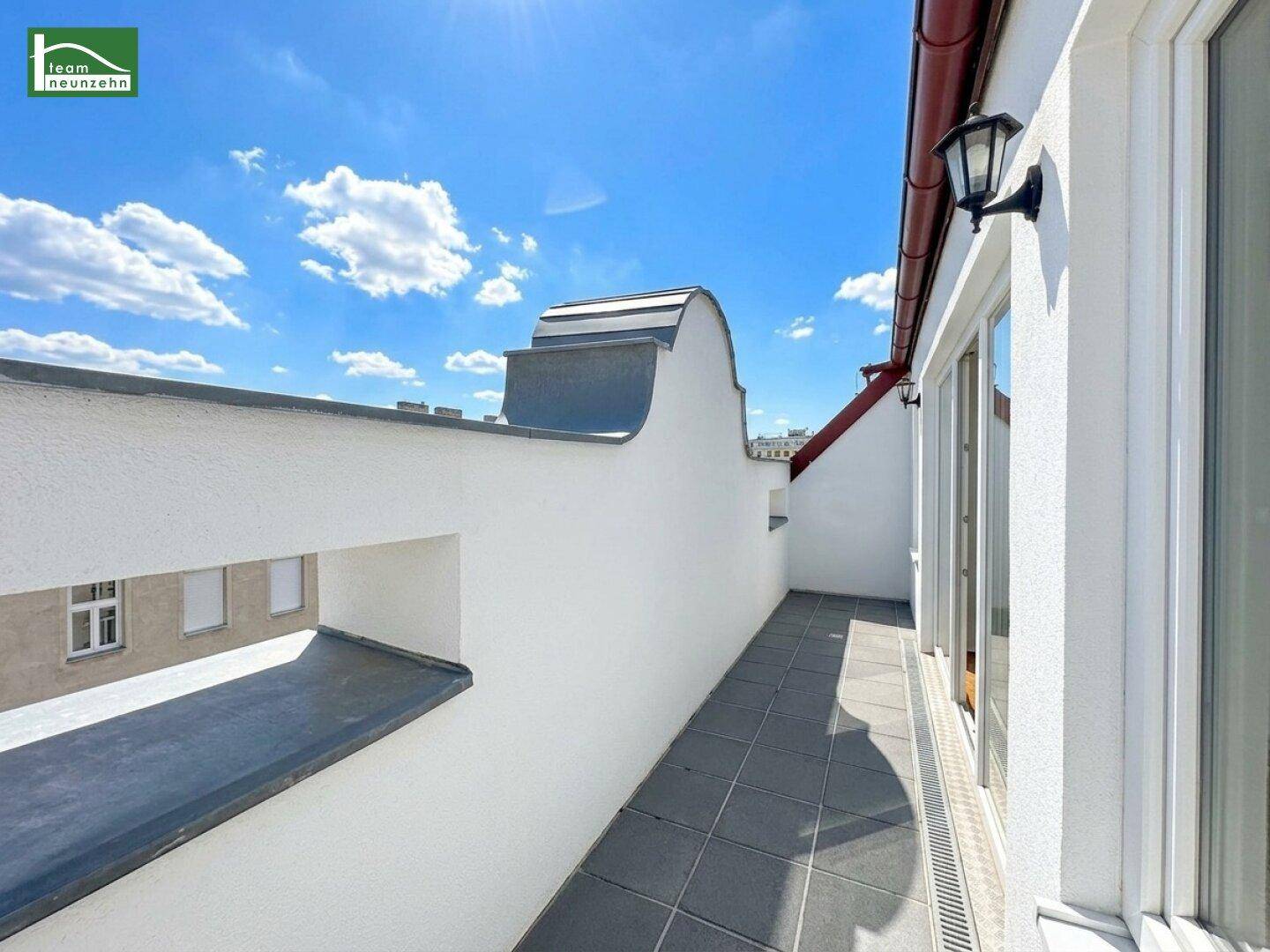 Sonniger Dachterrassenbereich mit Fliesenboden und hoher Brüstung unter blauem Himmel.
