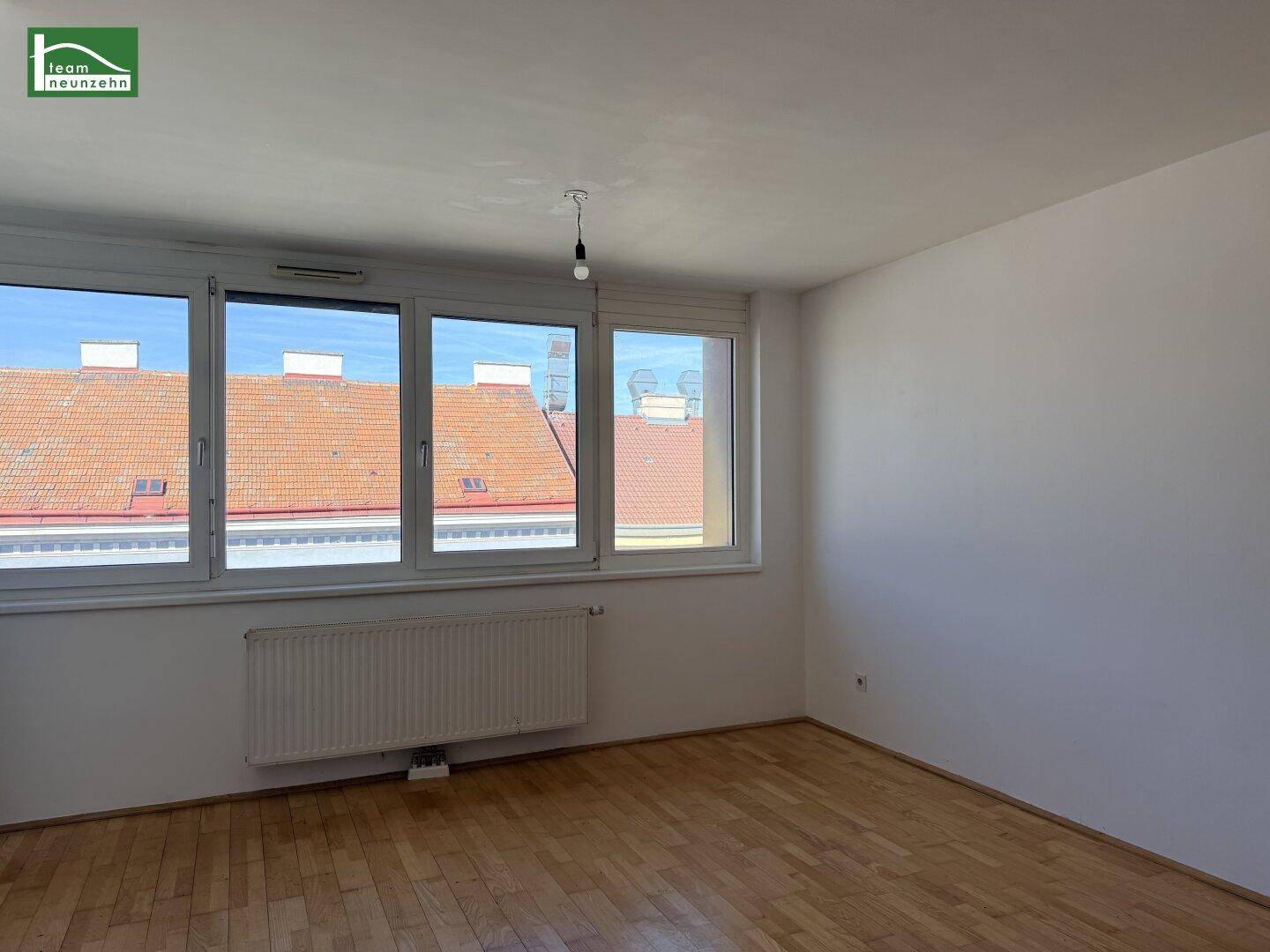 Leerer Raum mit großen Fenstern, die einen Blick auf die Dächer bieten, und einem Heizkörper.