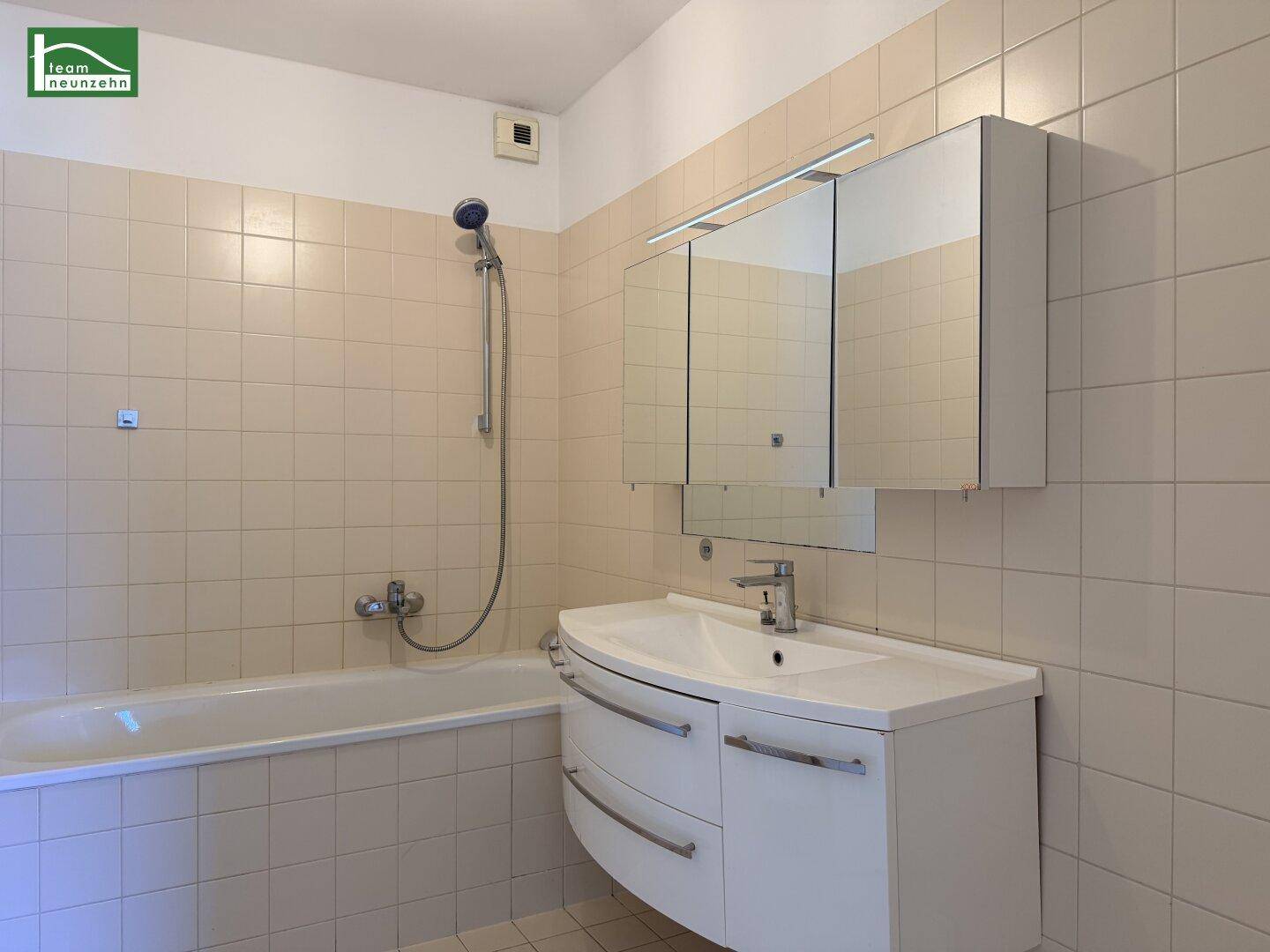 Badezimmer mit beigefarbenen Fliesen, Badewanne mit Duschfunktion und Waschtisch mit Spiegelschrank.