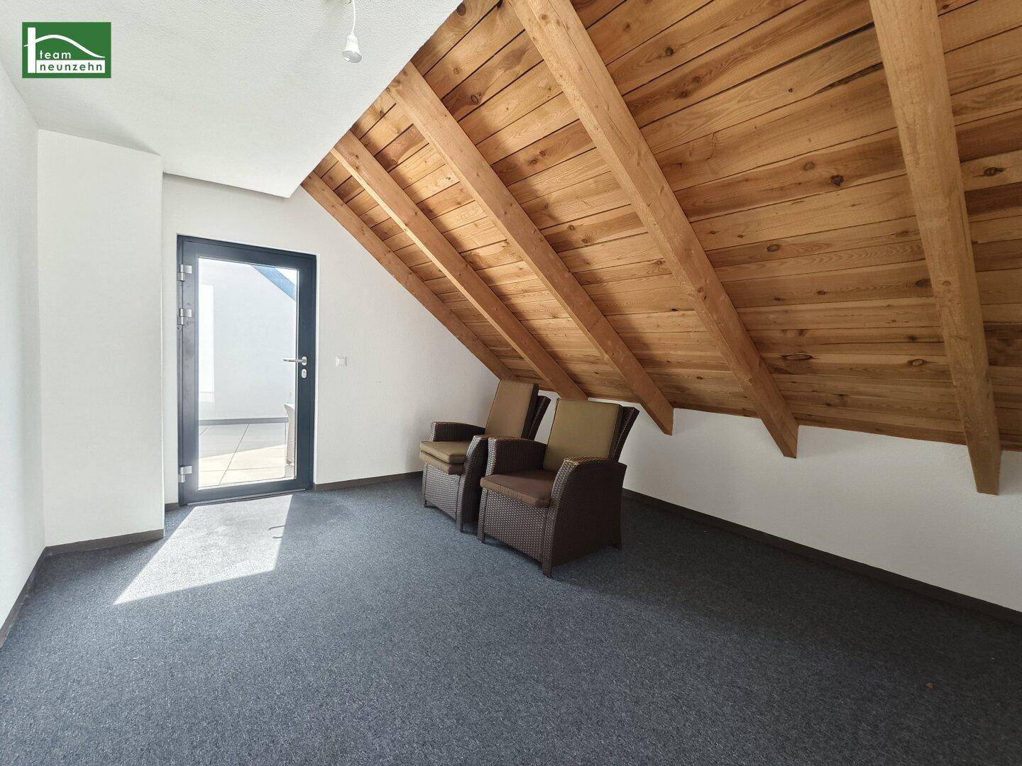 Dachzimmer mit sichtbaren Holzbalken, Teppichboden und Zugang zu einem Außenbereich.