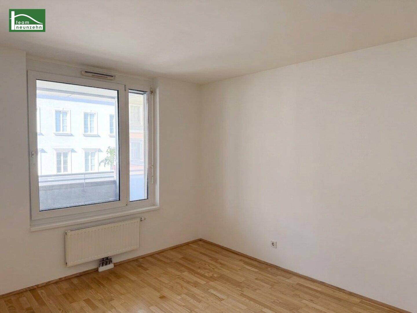 Helles Schlafzimmer mit Holzboden und Blick auf die umliegenden Gebäude durch das Fenster.