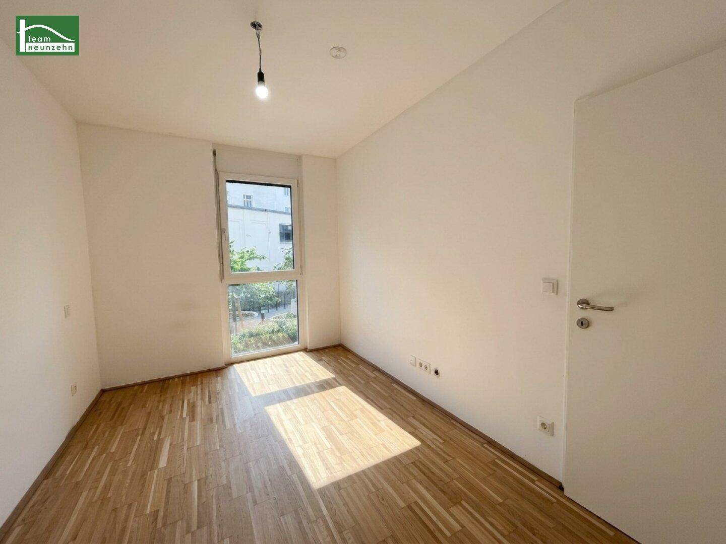 Unmöbliertes Schlafzimmer mit hellem Parkettboden und großem Fenster mit Blick ins Grüne.
