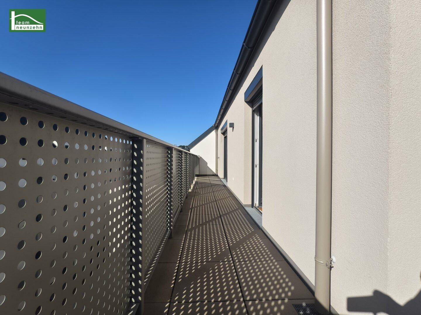 Langer Balkon mit perforiertem Metallgeländer und Fliesenboden unter blauem Himmel.