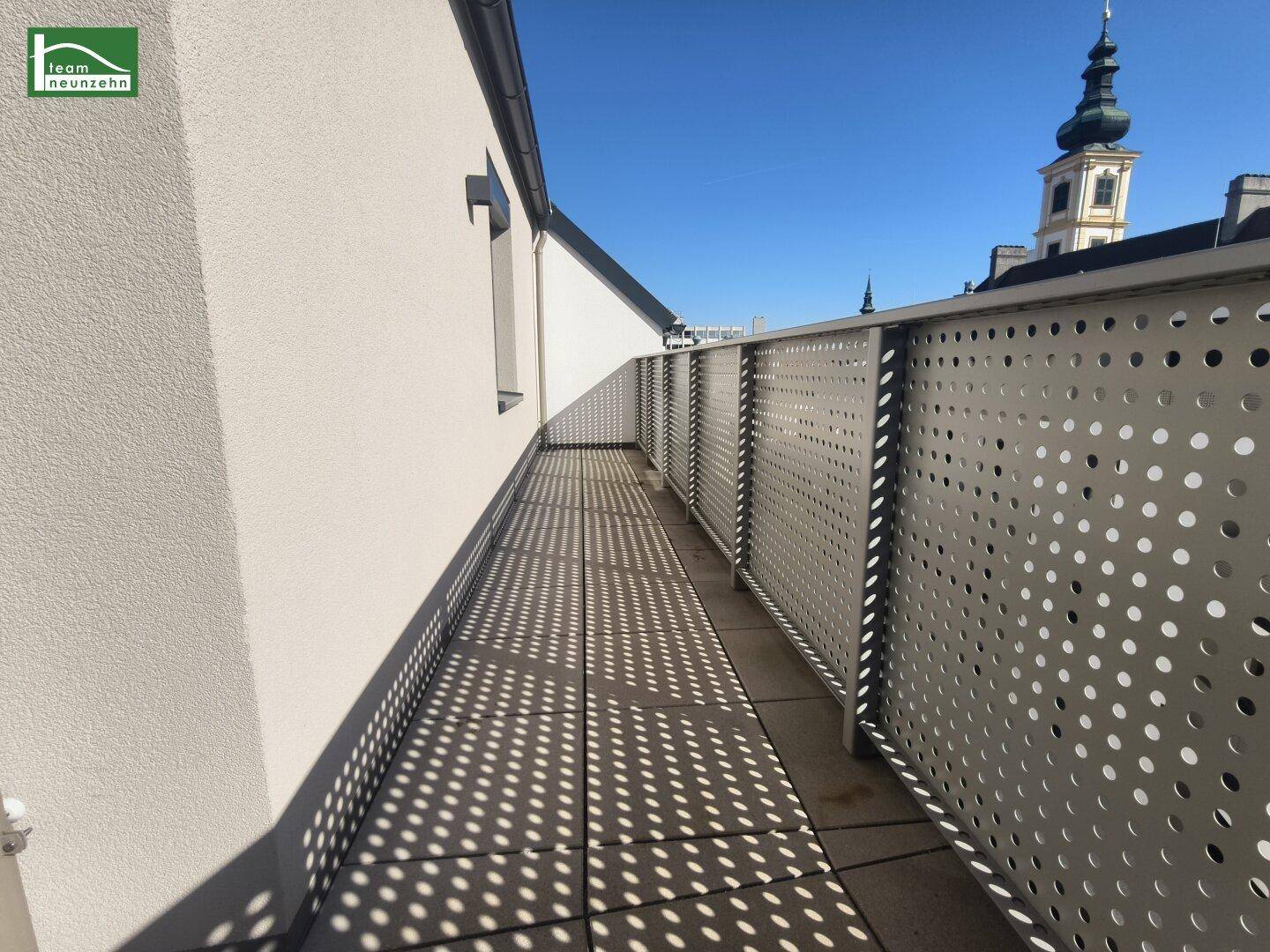 Sonniger Balkon mit modernem Geländer und Blick auf einen Kirchturm in der Ferne.
