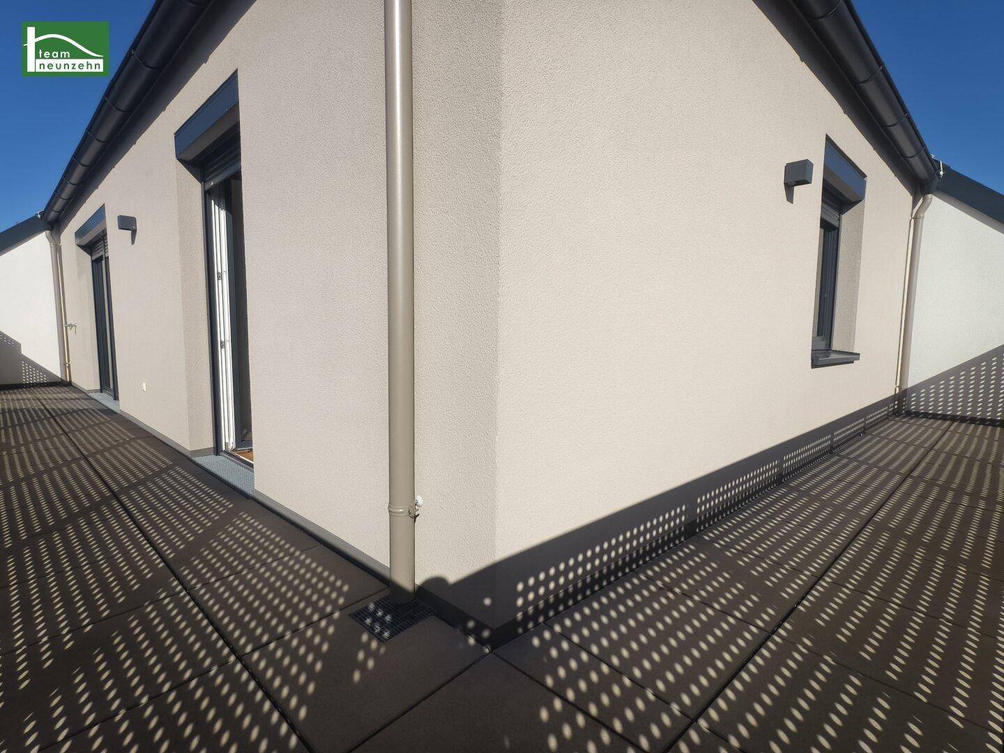 Eckbereich einer modernen Dachterrasse mit hellem Fassadenputz und Rollläden an den Fenstern.