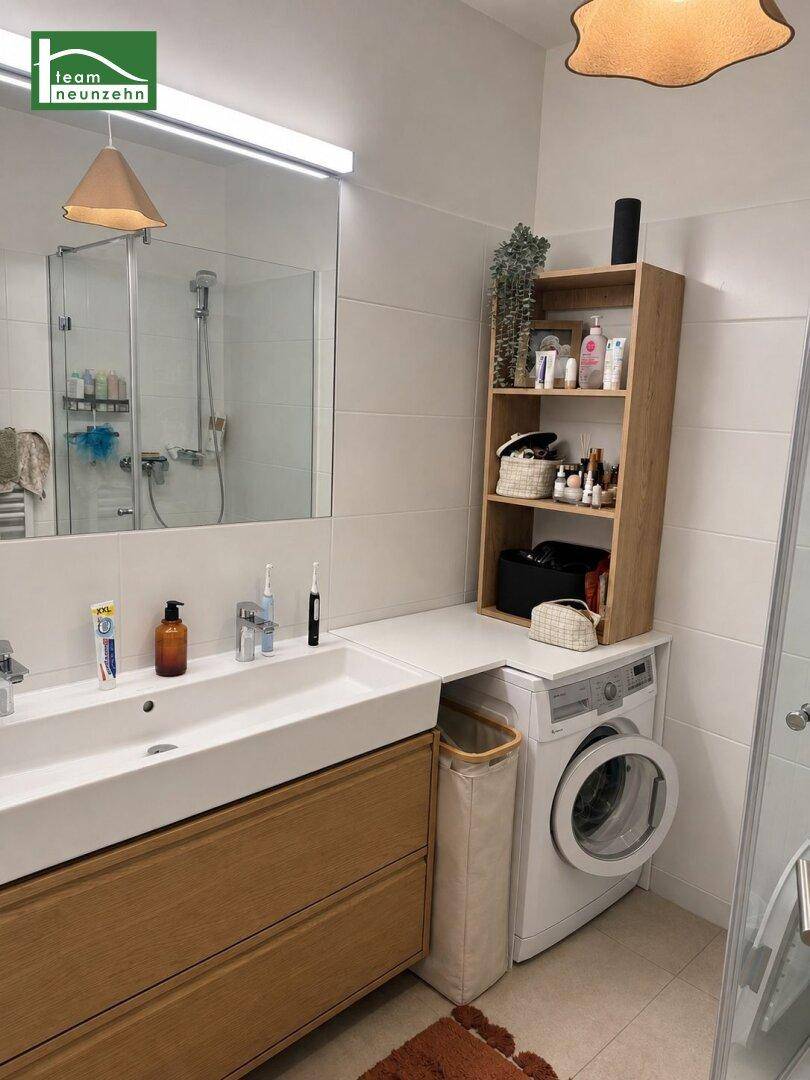 Modernes Badezimmer mit Doppelwaschbecken, Dusche und integrierter Waschmaschine unter einer Ablage.