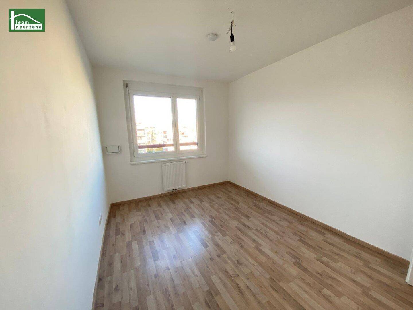 Helles, leeres Zimmer mit Laminatboden und einem Fenster, das viel Tageslicht hereinlässt.