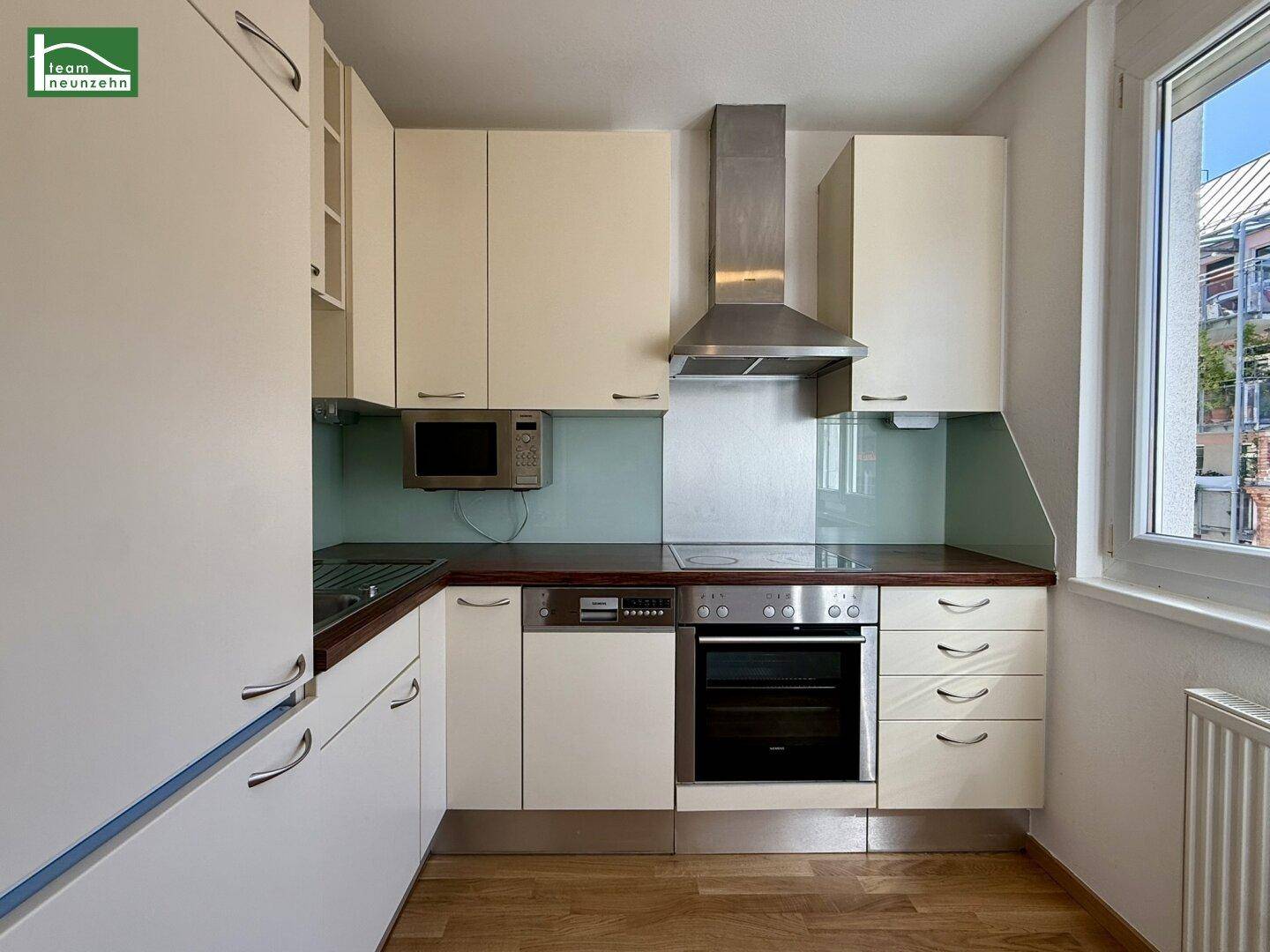 Moderne Einbauküche mit beigen Hochglanzschränken, Edelstahlgeräten und einem Fenster mit Blick nach draußen.