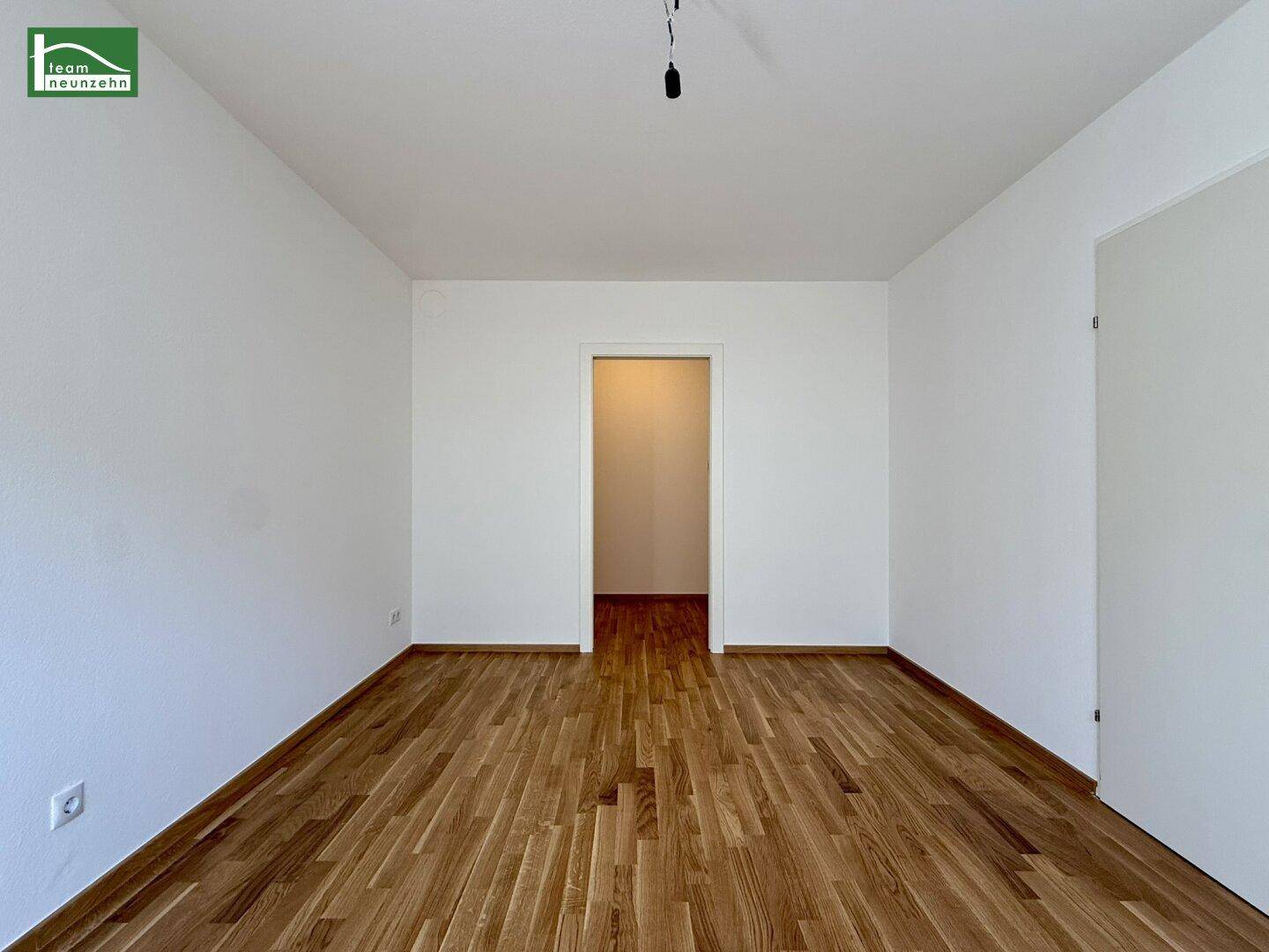 Geräumiger, leerer Wohnraum mit Holzparkettboden und Zugang zu einem weiteren Zimmer.