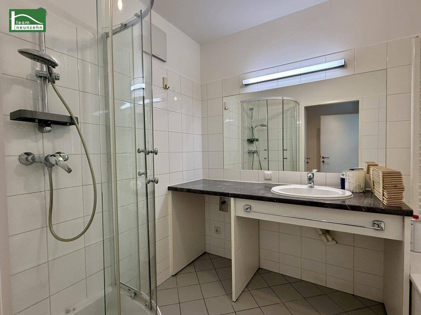 Badezimmer mit Duschkabine, Waschtisch mit dunkler Platte und weißen Wandfliesen.