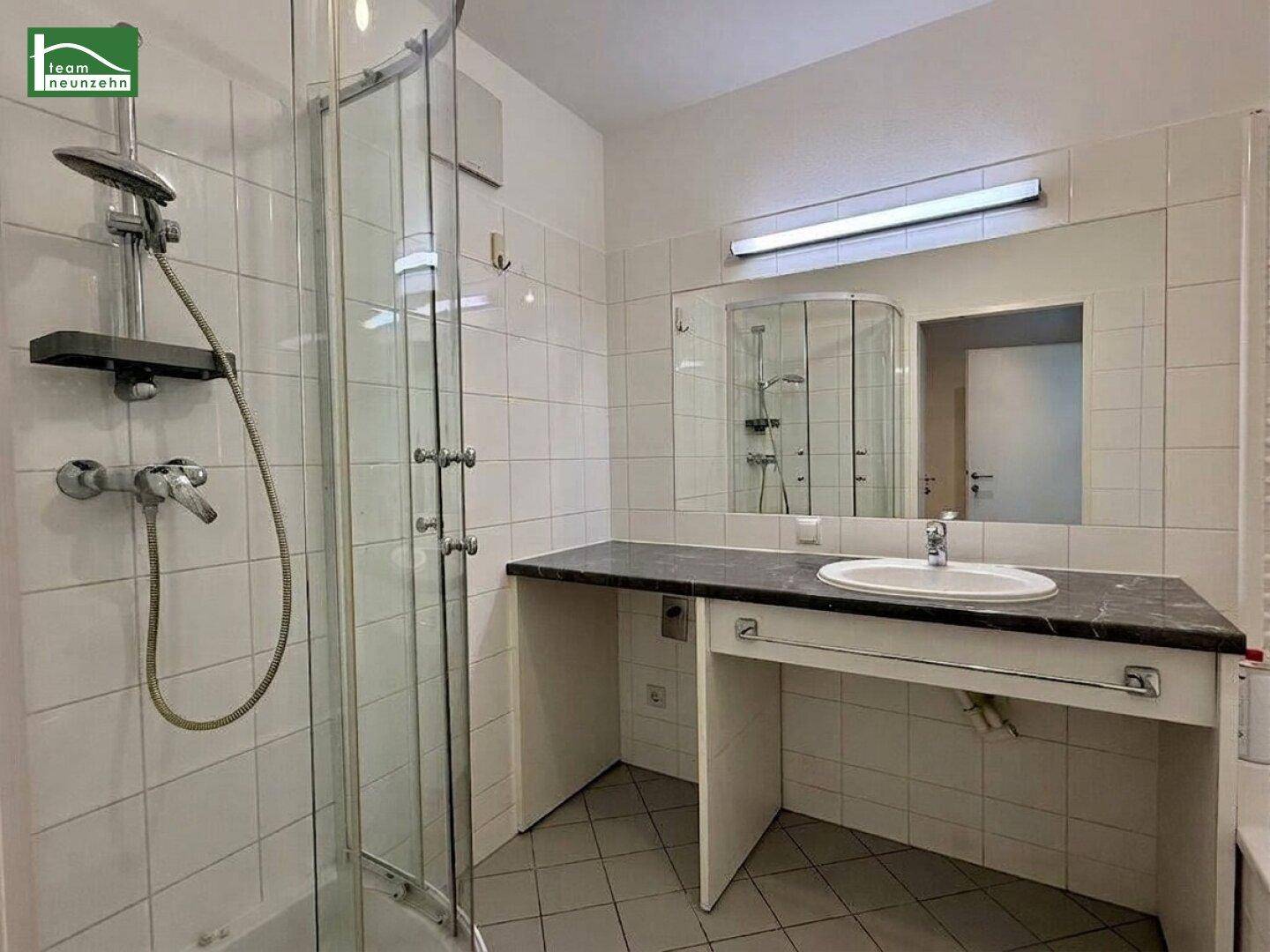 Badezimmer mit Duschkabine, Waschbecken mit Ablage und großen weißen Wandfliesen, in gutem und gepflegtem Zustand.