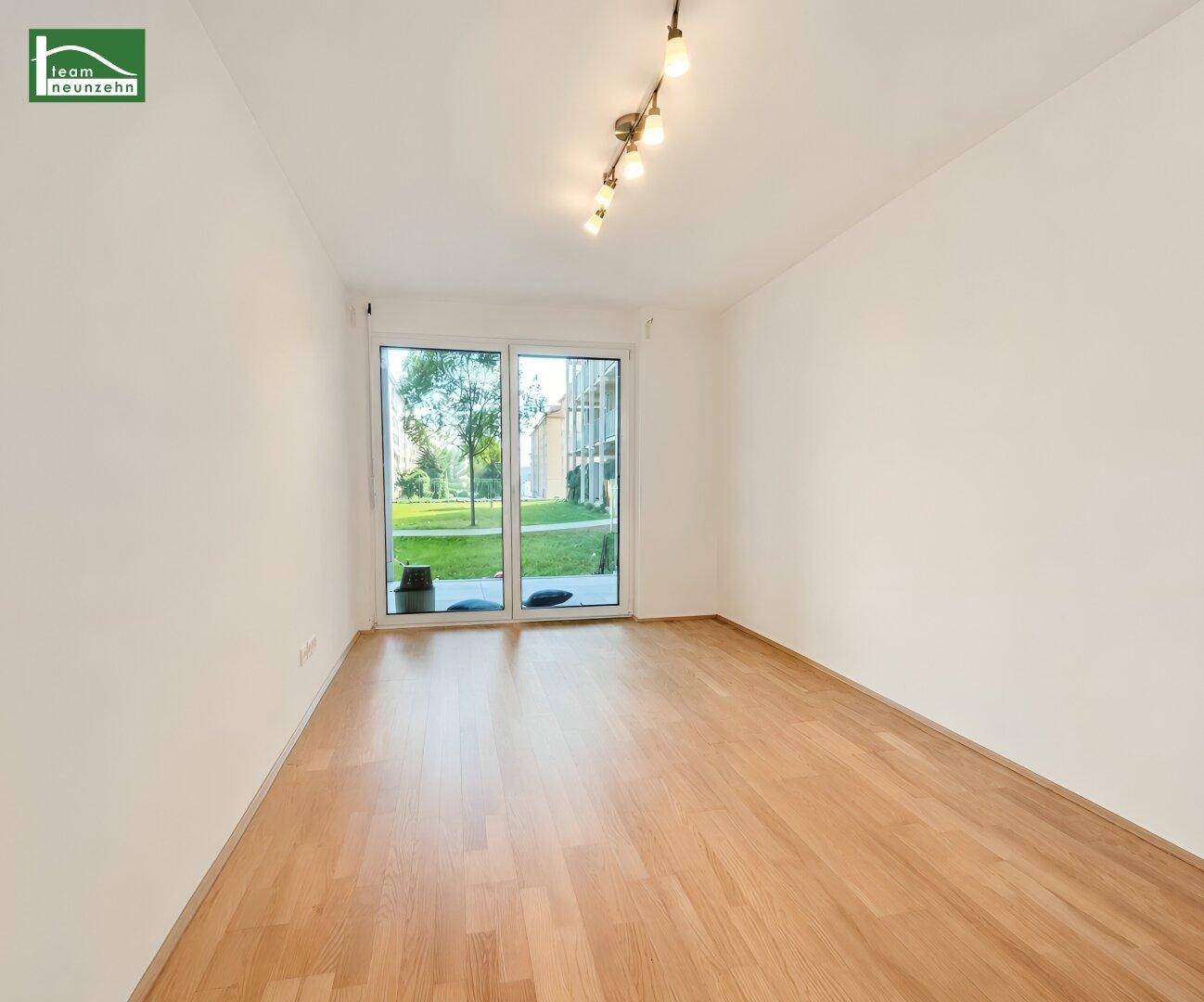 Leeres Zimmer mit Holzboden und großen Fenstern mit Blick ins Grüne.