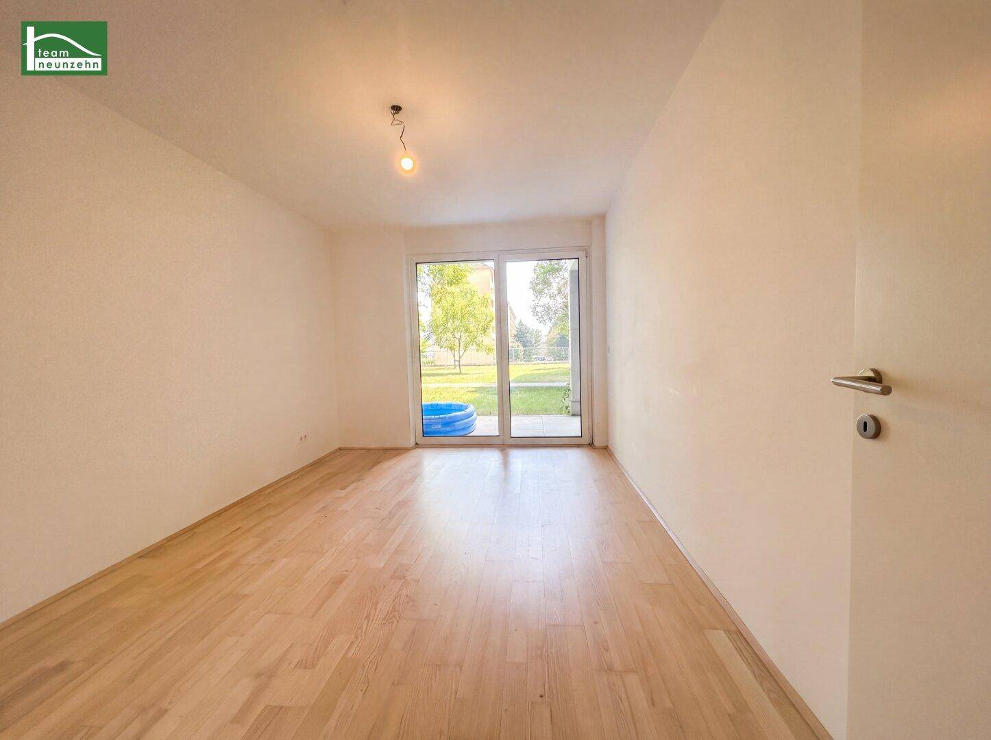Geräumiges, leeres Zimmer mit Holzboden und großen Fenstern zum Garten.