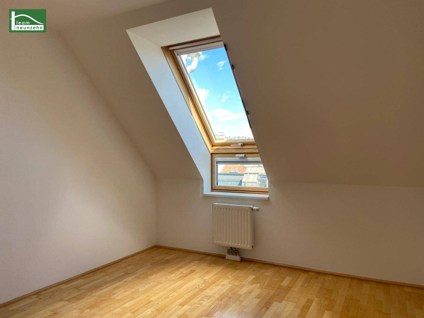 Heller Raum mit schrägen Wänden, Parkettboden und einem großen Dachfenster mit Blick nach draußen.