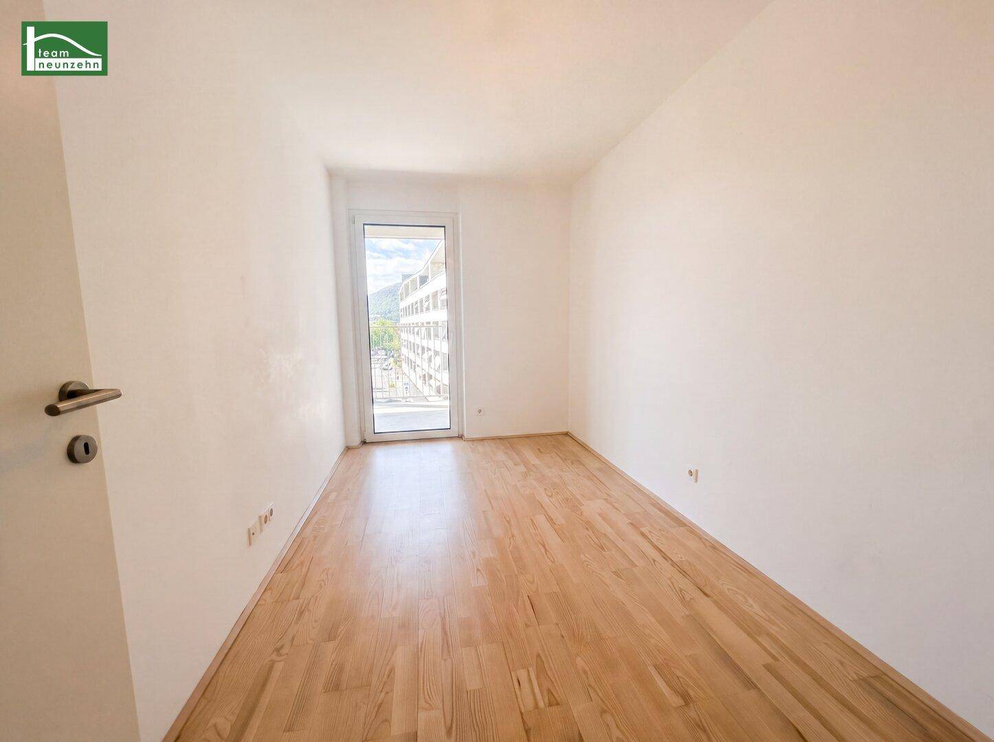 Leeres Schlafzimmer mit Parkettboden und einer Tür, die zu einem französischen Balkon führt.