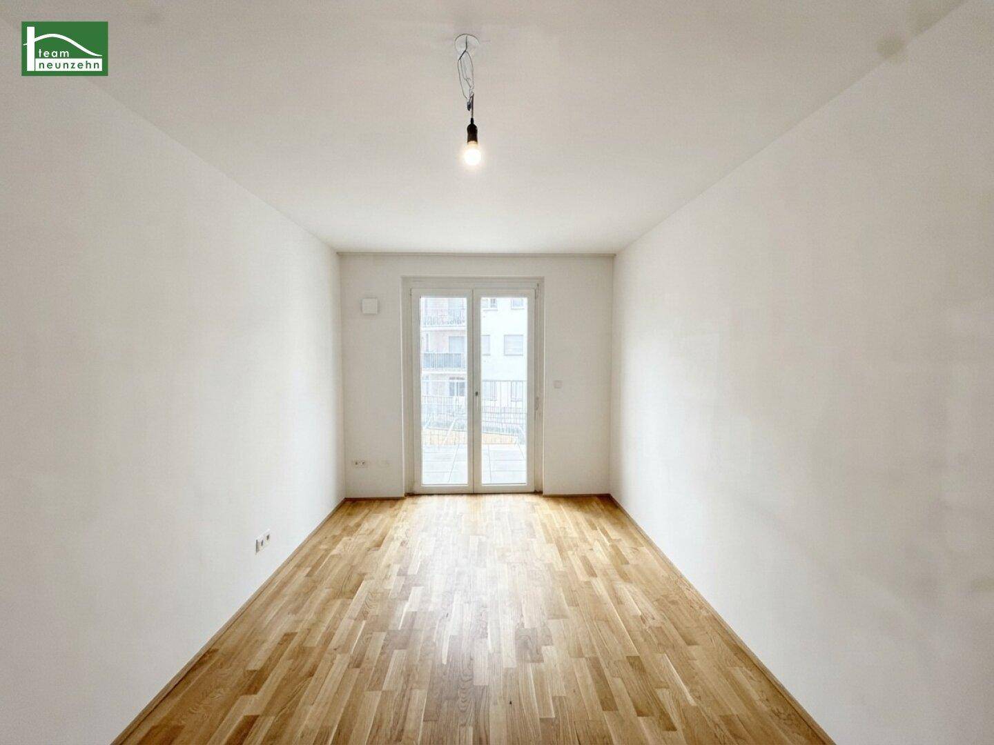 Leeres Zimmer mit hellem Holzboden und großen Fenstertüren, die zu einem Balkon führen.