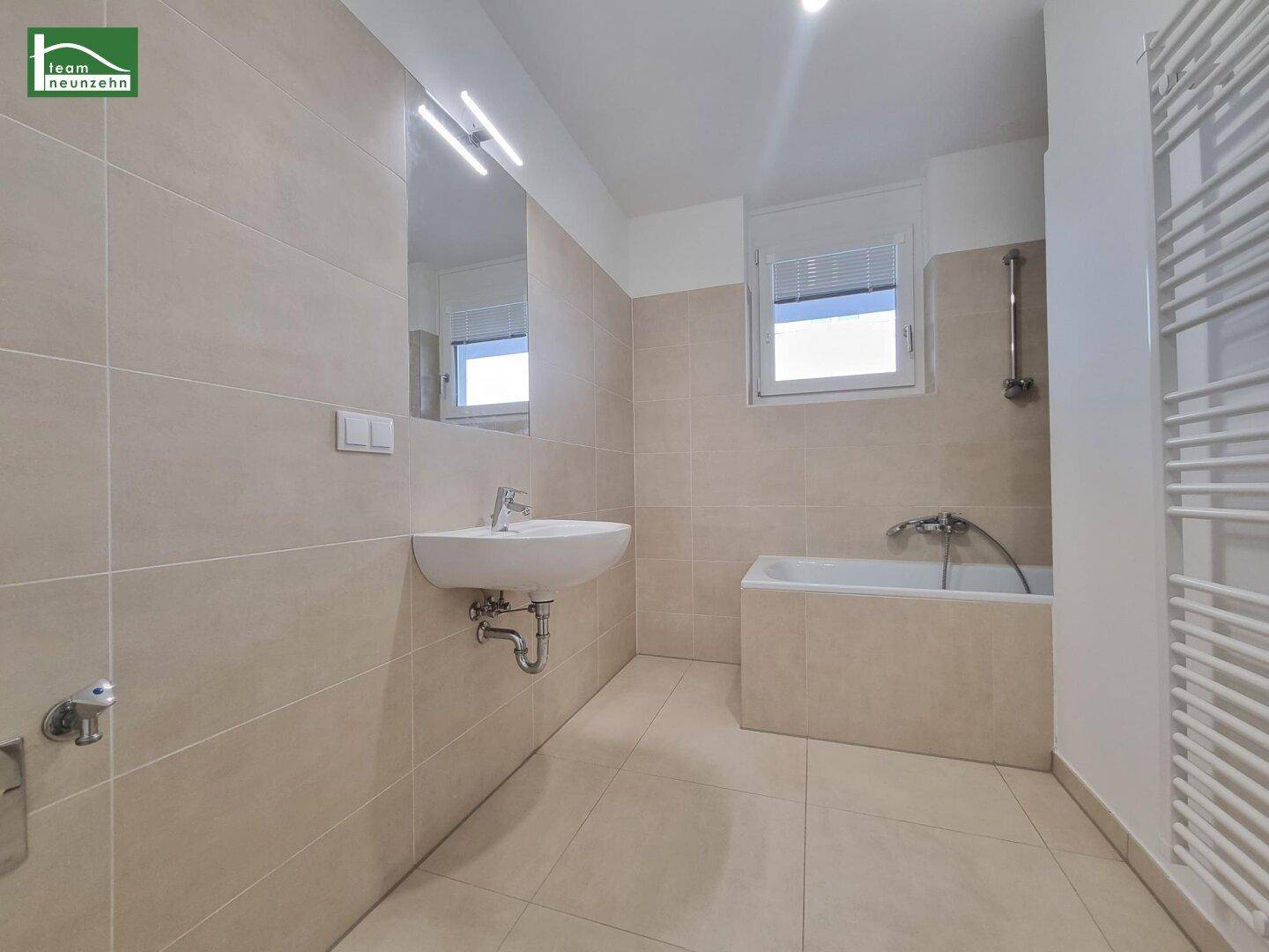 Helles Badezimmer mit Badewanne, Einzelwaschbecken, beheiztem Handtuchhalter und Fenster.