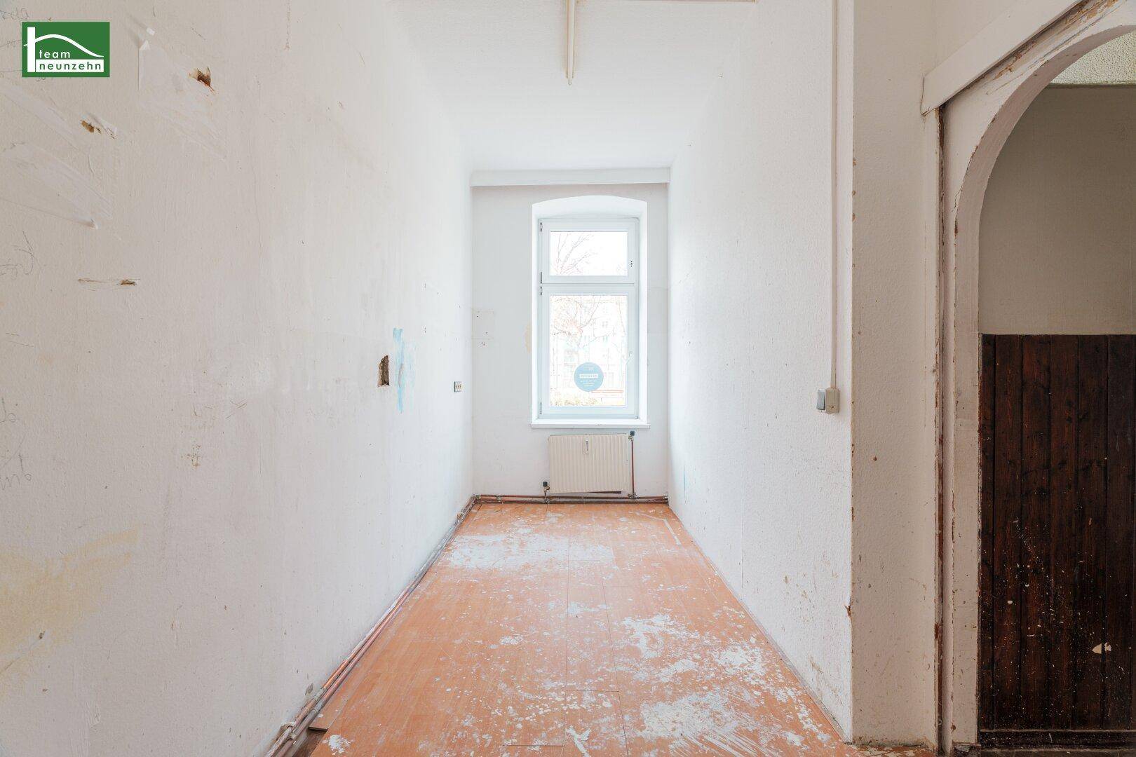 Schmaler Raum mit einem Fenster, abgenutztem Boden und einer Holztür.