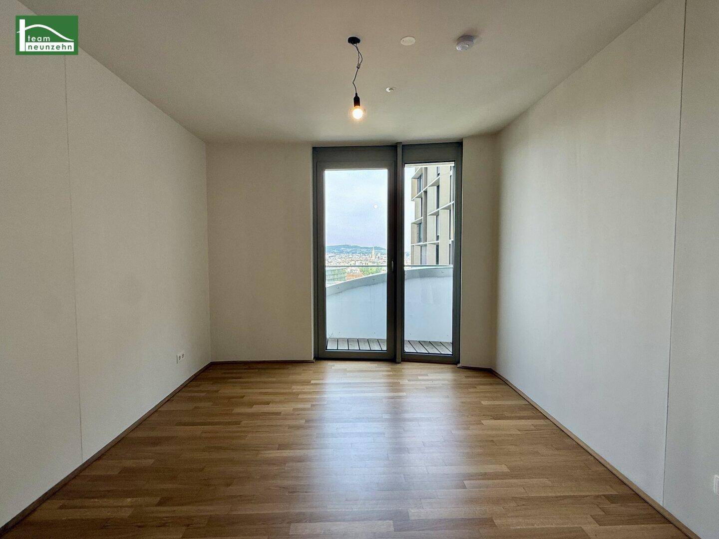 Leeres Zimmer mit Parkettboden und großem Fenster, das Zugang zu einem Balkon bietet.