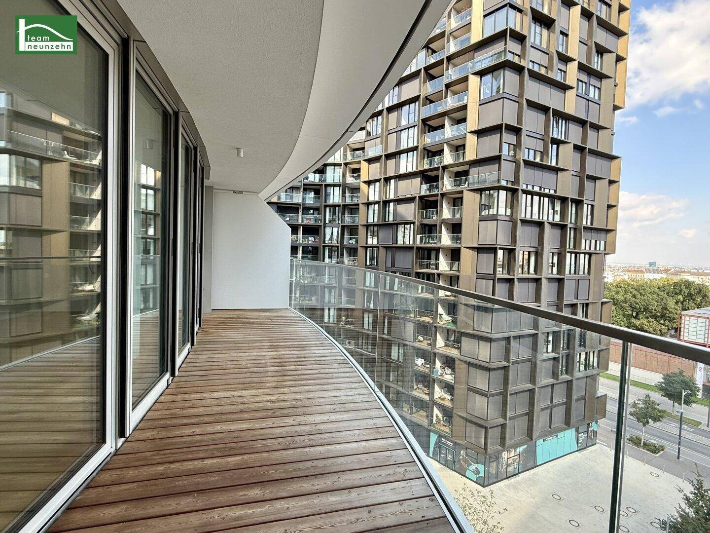 Geschwungener Balkon mit Holzbelag und Glasgeländer, bietet Blick auf moderne Architektur.