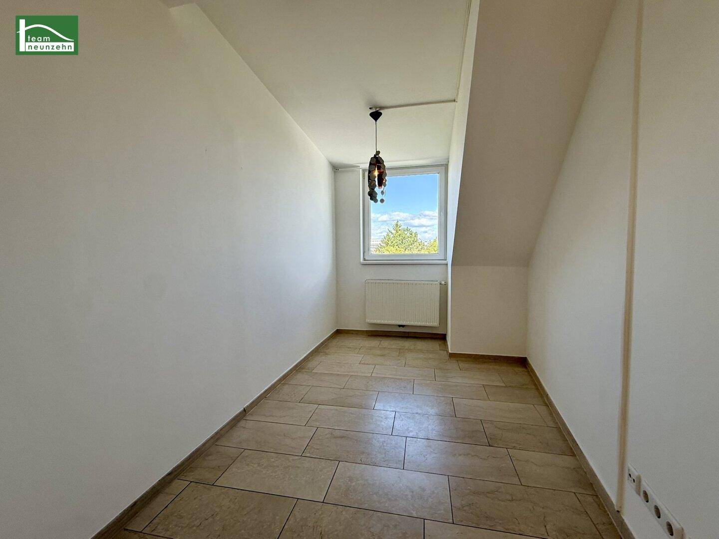 Kleines Zimmer mit Fliesenboden, Fenster und Heizkörper unter der Dachschräge.
