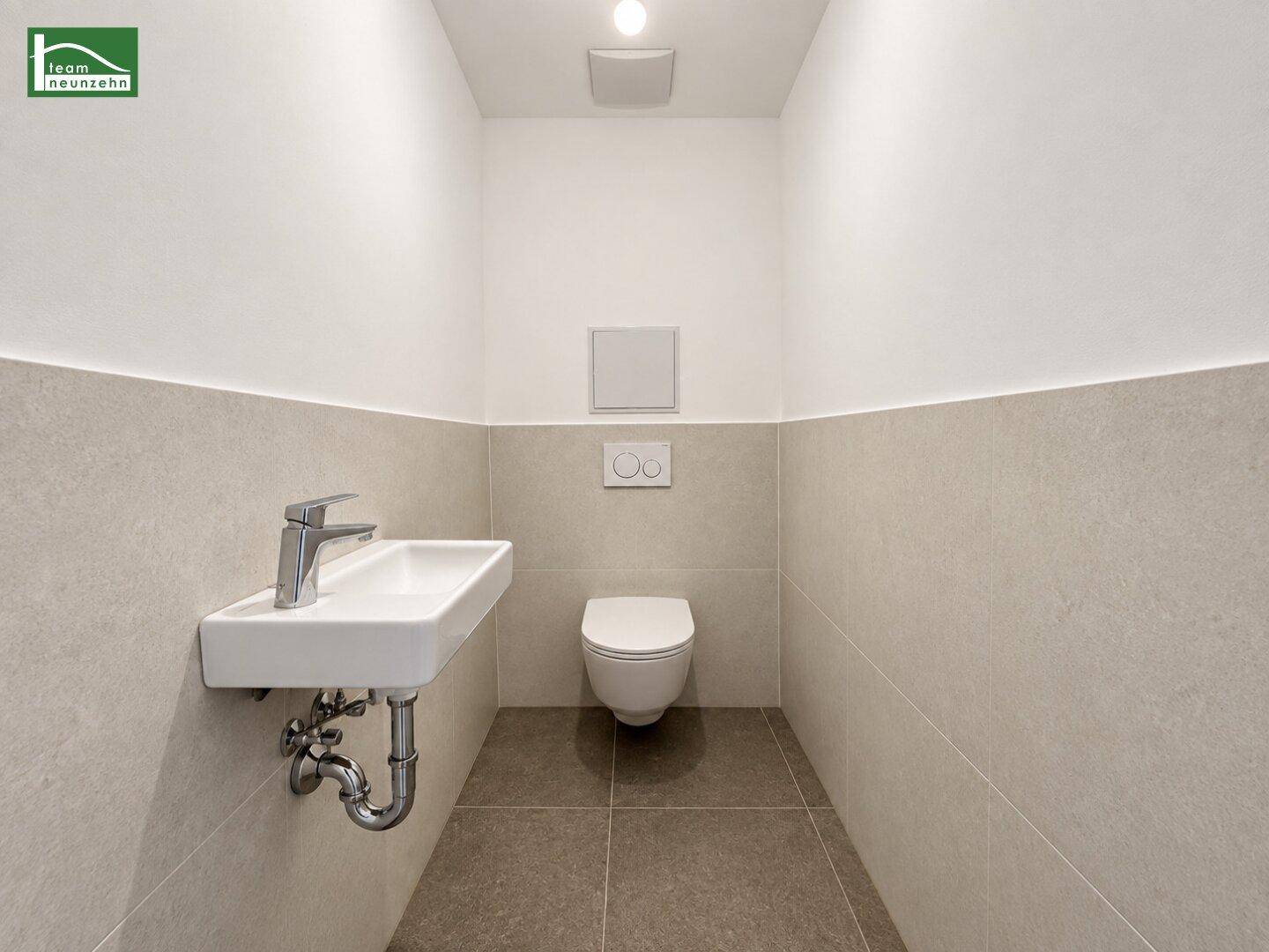 Unmöbliertes Gäste-WC mit Wand-WC und kleinem Waschbecken, hell gefliest.