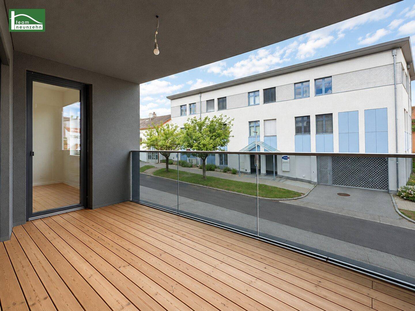 Geräumiger Balkon mit Holzboden und Glasgeländer, bietet einen Blick auf die umliegenden Gebäude.