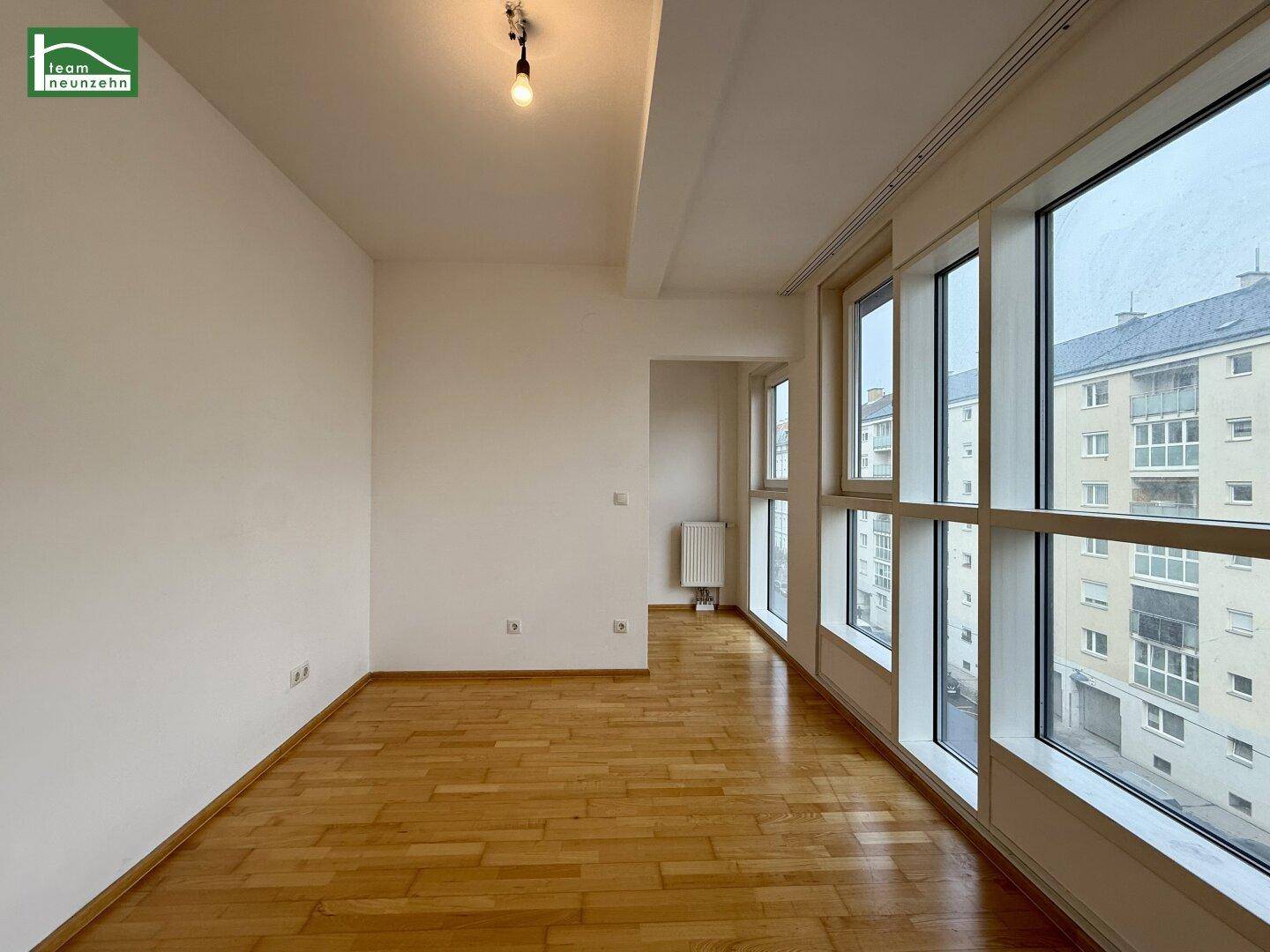 Geräumiges Zimmer mit Parkettboden und einer Fensterfront, die einen weiten Blick über die Stadt bietet.