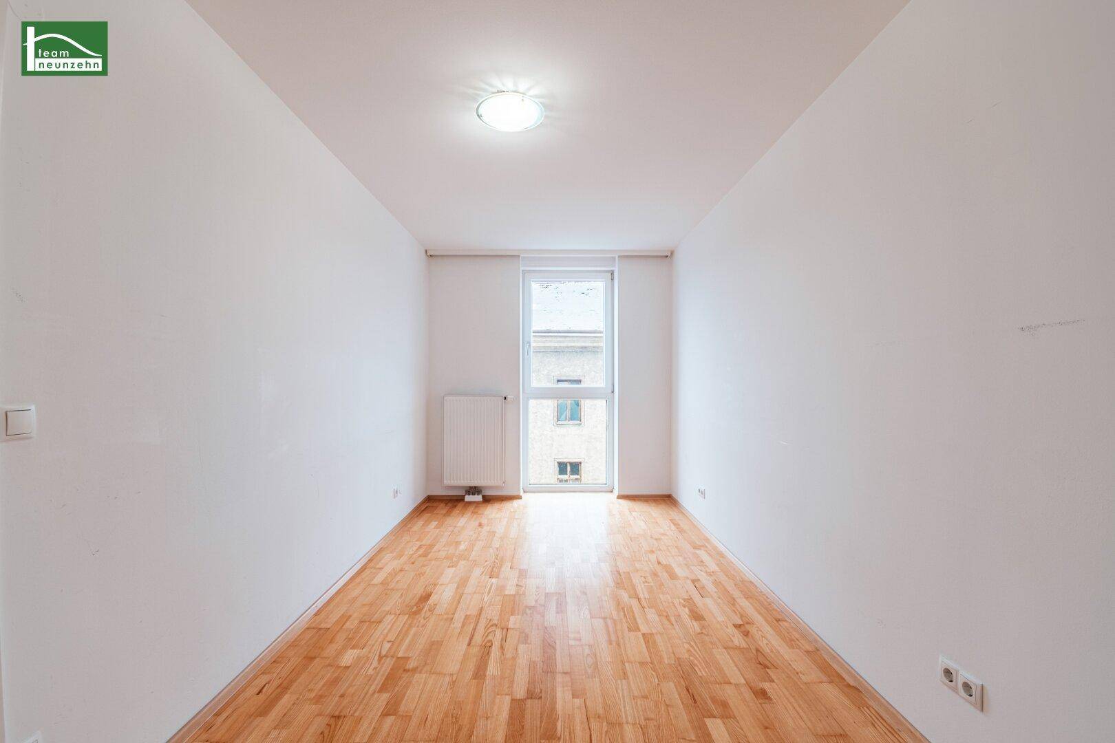 Heller, leerer Raum mit Parkettboden und einem Fenster, das Tageslicht hereinlässt.
