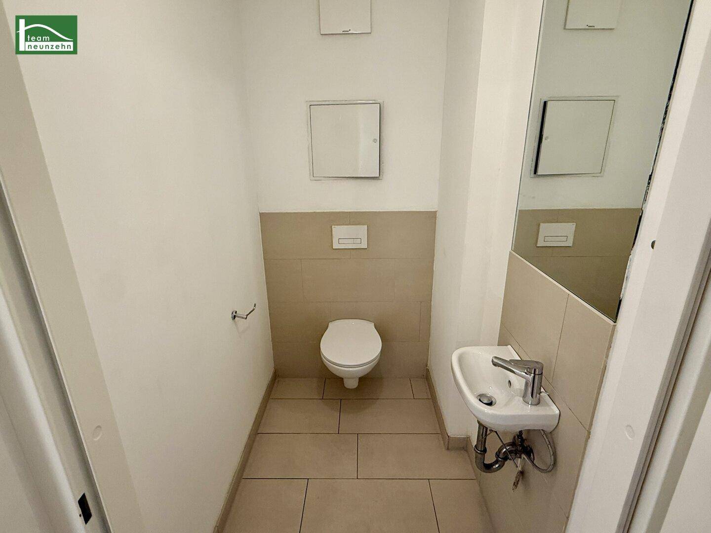 Separates WC mit Wand-WC, kleinem Waschbecken und hellen Fliesen.