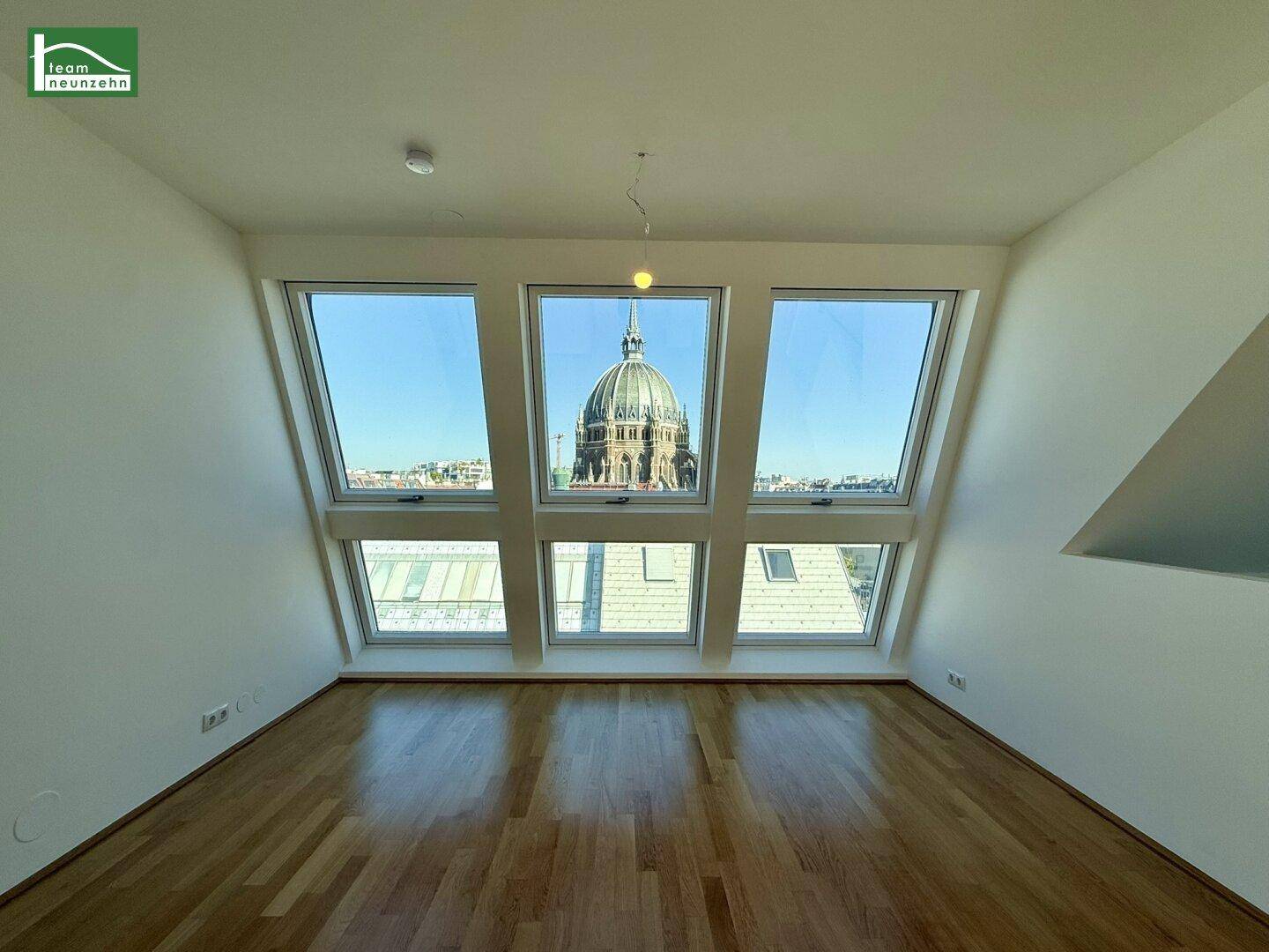 Heller Raum im Dachgeschoss mit schrägen Fenstern, die einen beeindruckenden Blick auf eine historische Kuppel ermöglichen.