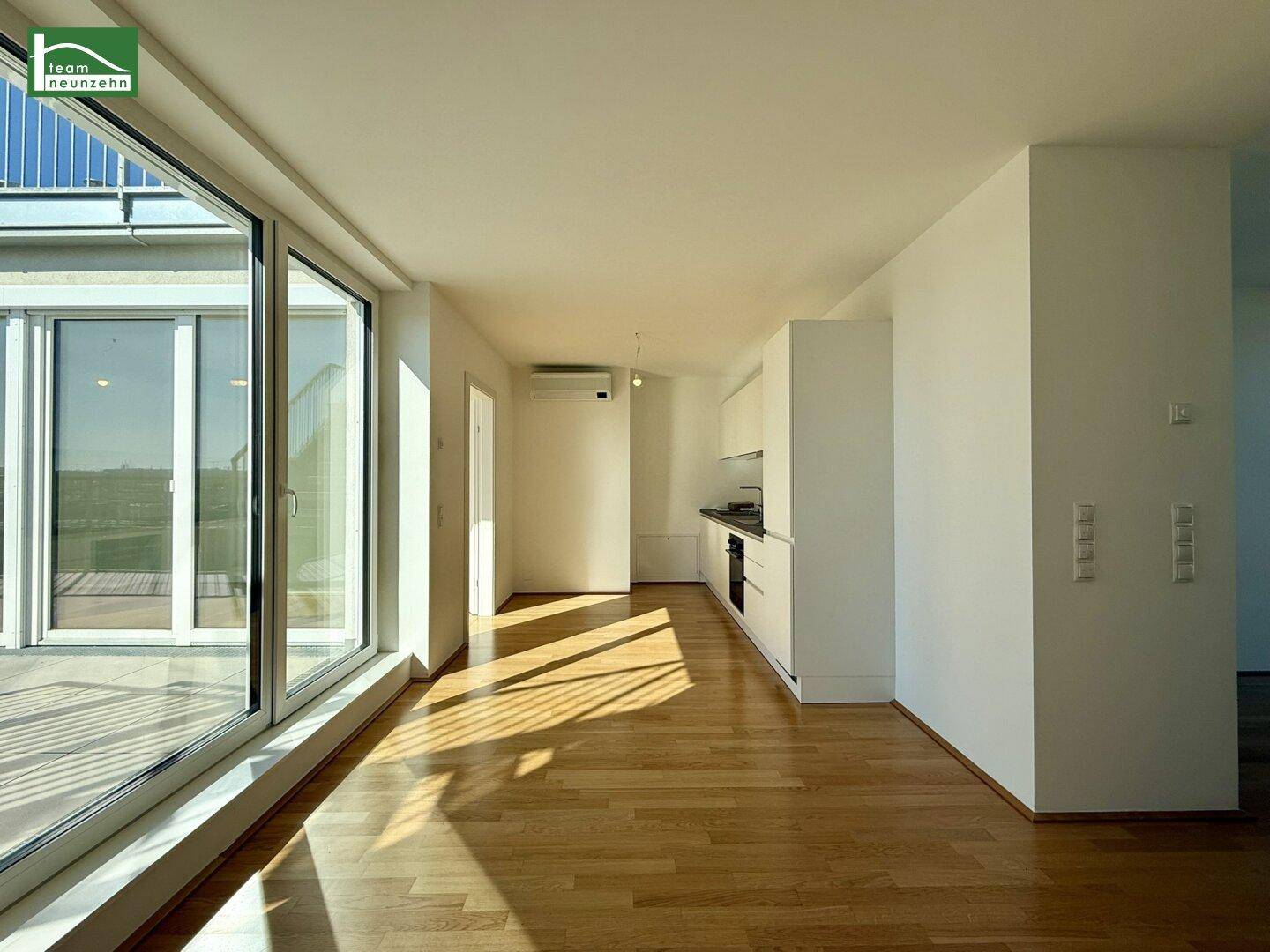 Heller Wohnbereich mit Holzboden und großen Fenstern, die viel Sonnenlicht hereinlassen und eine kleine Küchenzeile zeigen.
