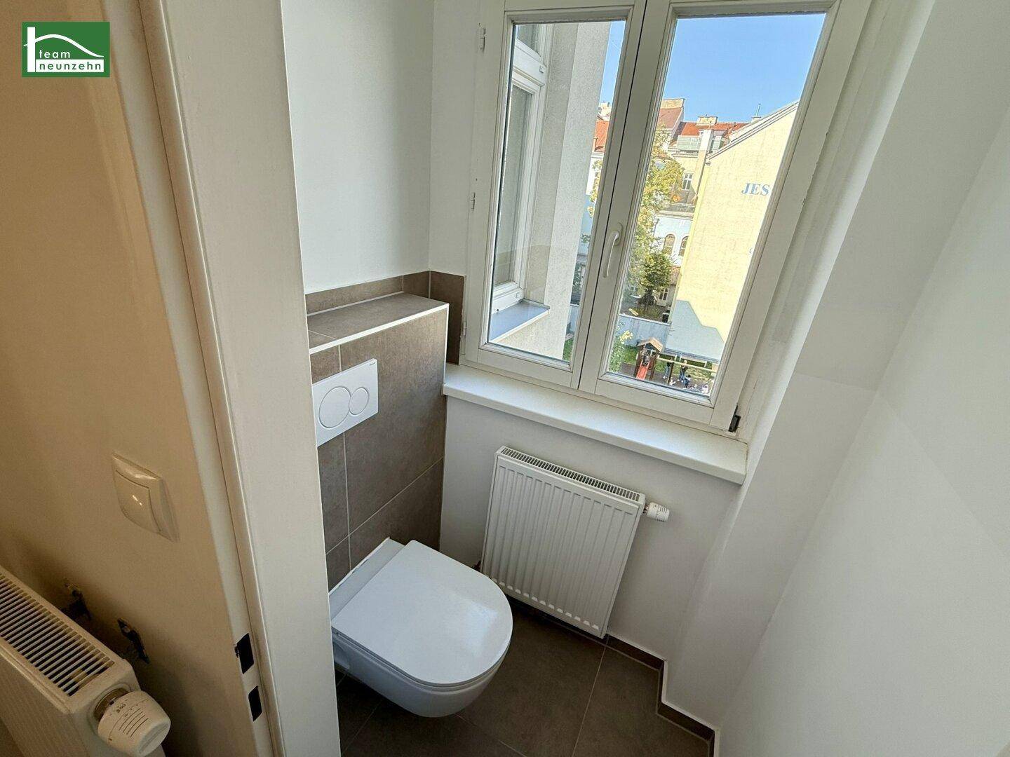 Separates WC mit wandhängender Toilette, kleinem Waschbecken und Fenster, hell gefliest.