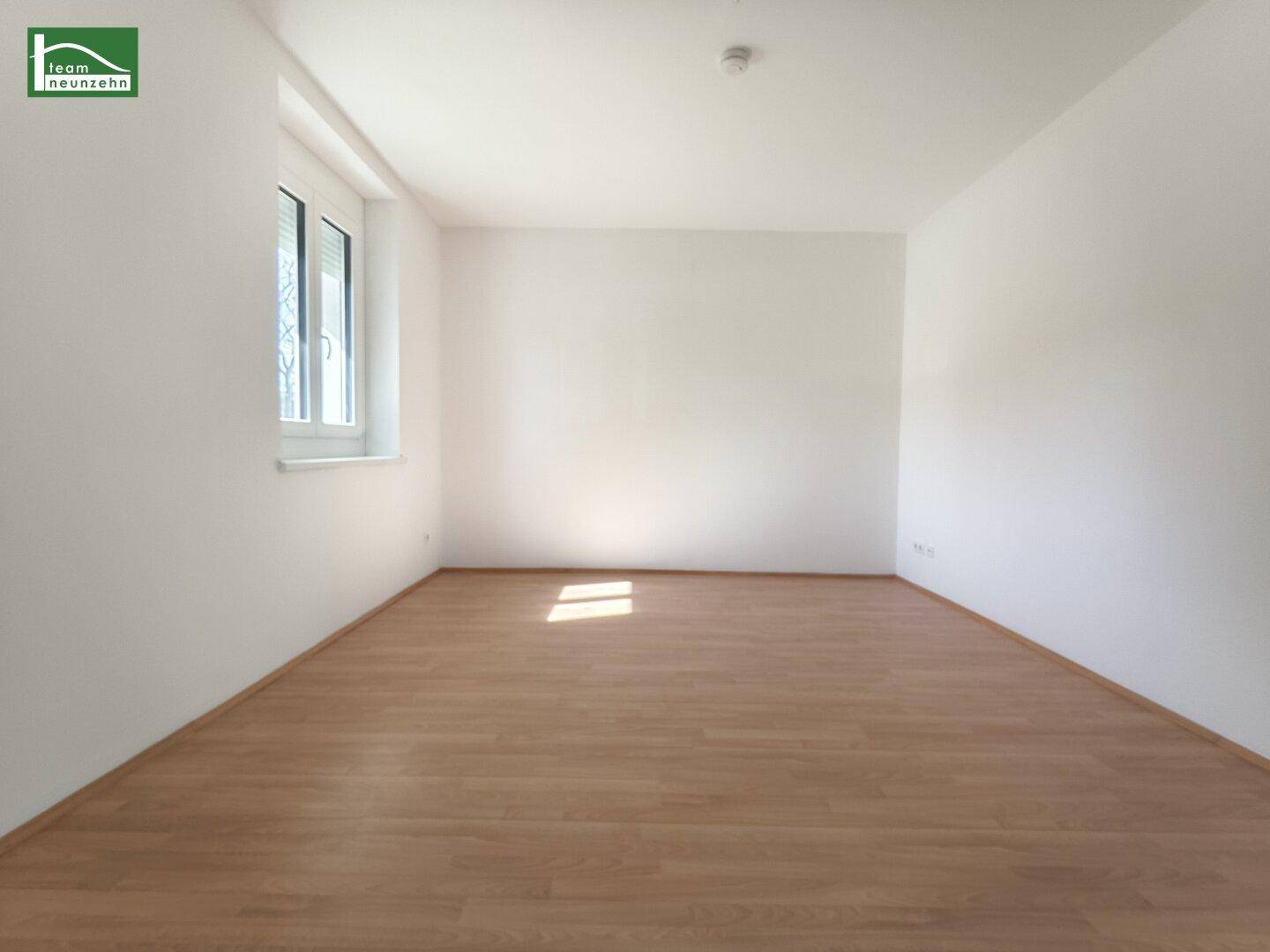 Lichtdurchflutetes Zimmer mit Laminatboden und einem Fenster mit Rollladen, das viel Tageslicht hereinlässt.