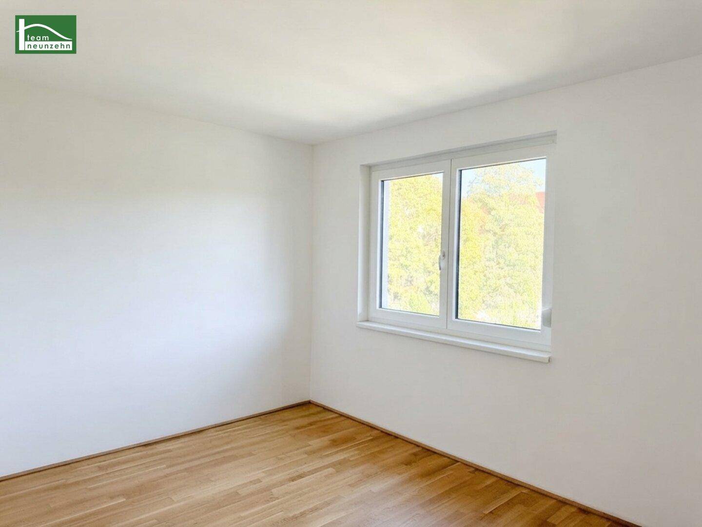 Helles Schlafzimmer mit Parkettboden und einem Fenster mit Blick auf Bäume.