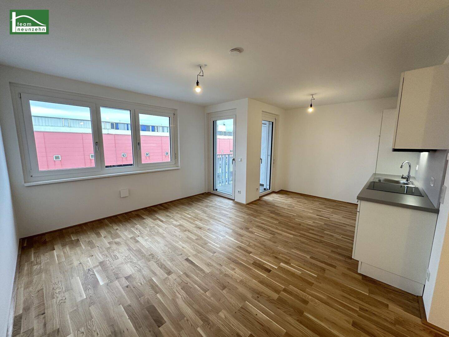 Heller offener Wohnbereich mit Holzboden, großen Fenstern und Zugang zu einem Balkon.