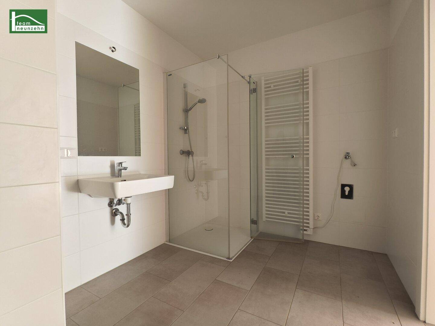 Modernes Badezimmer mit ebenerdiger Dusche, Waschbecken und beheiztem Handtuchhalter, in hellen Fliesen gehalten.