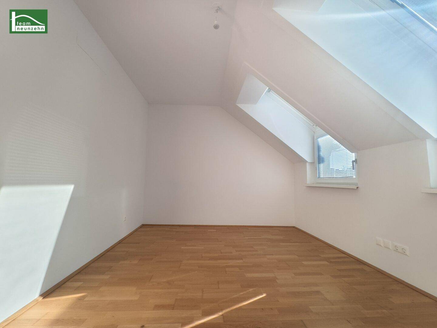 Helles Dachzimmer mit schrägen Wänden und Dachfenstern, ausgestattet mit einem warmen Holzparkettboden.