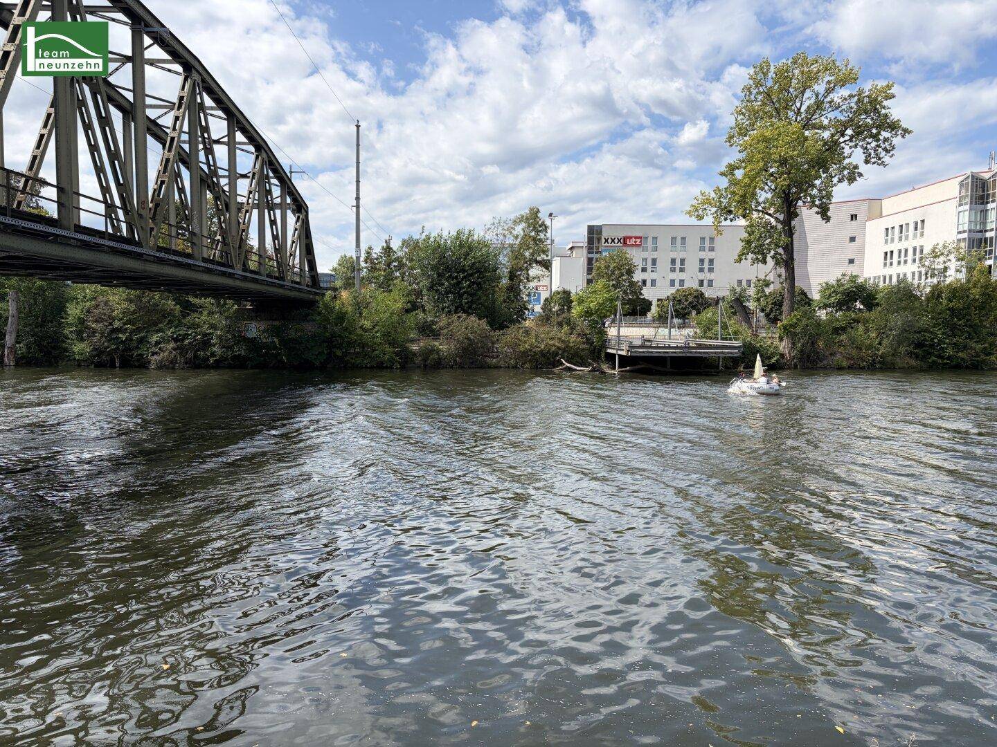 Flusslandschaft mit einer Eisenbahnbrücke und Gebäuden am Ufer unter einem sonnigen Himmel.