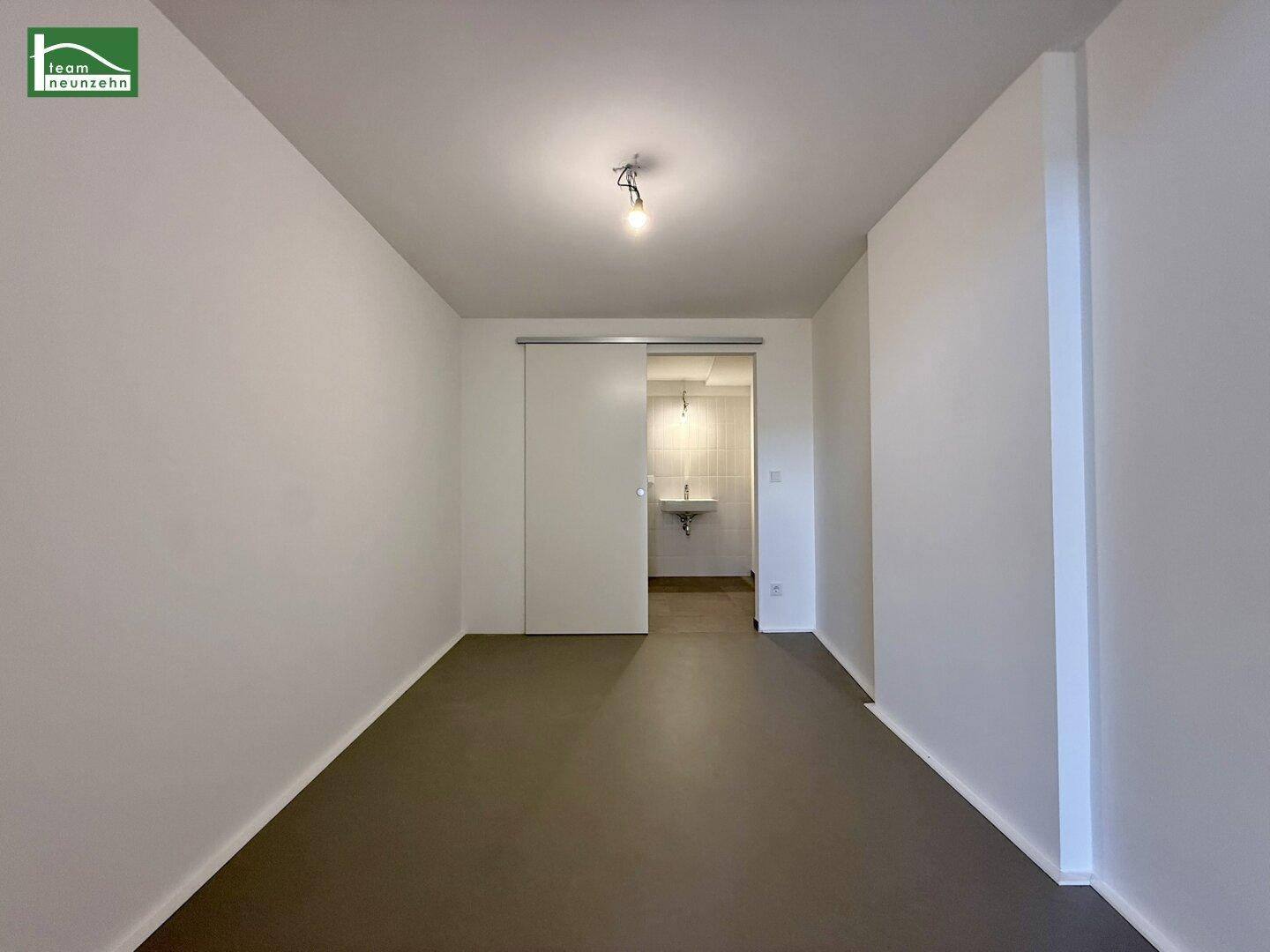 Heller Raum mit grauem Boden und Schiebetür, die zu einem Badezimmer führt.