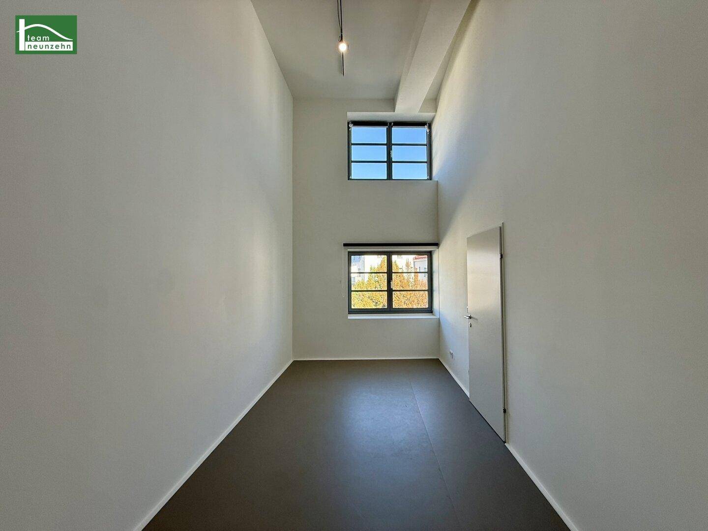 Heller Raum mit hohen Decken, zwei Fenstern und grauem Boden, vielseitig nutzbar.