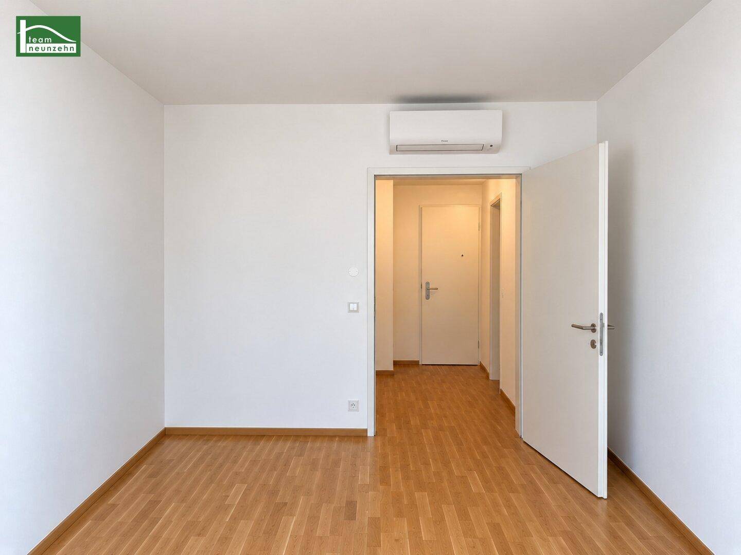 Heller, leerer Raum mit Holzboden, Klimaanlage und offenem Zugang zu einem Flur.