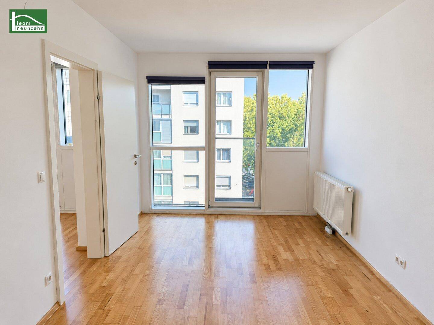 Geräumiges Zimmer mit Parkettboden, großen Fenstern und Blick auf die Umgebung mit Bäumen.